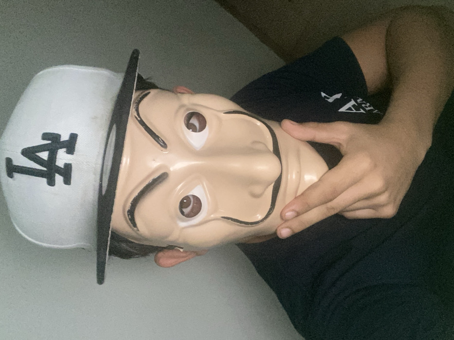A person wearing a plastic mask that resembles a cartoon man with thick eyebrows and a mustache is visible. They are also wearing a white "LA" baseball cap turned to the side. The person's hand is visible, touching the mask near the chin. The individual is wearing a dark blue shirt. The background appears to be a plain wall. The image is taken from a low angle.