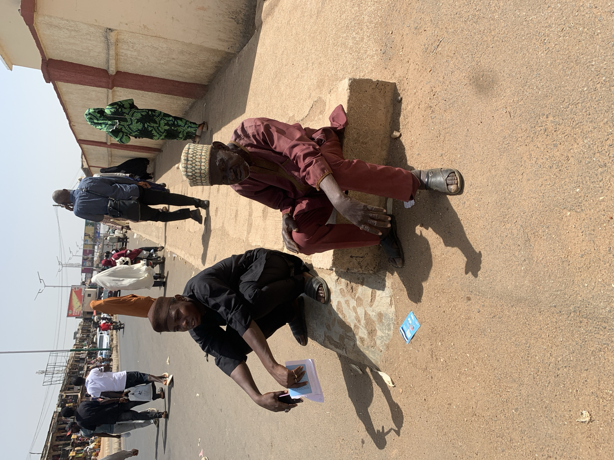 Two men are seated on the ground in Jos, Nigeria. One man, wearing a maroon outfit and a patterned cap, is leaning against a concrete structure. The other man is wearing black and is holding papers. A street scene with pedestrians and motorbikes is in the background. There is a building adjacent to the men.