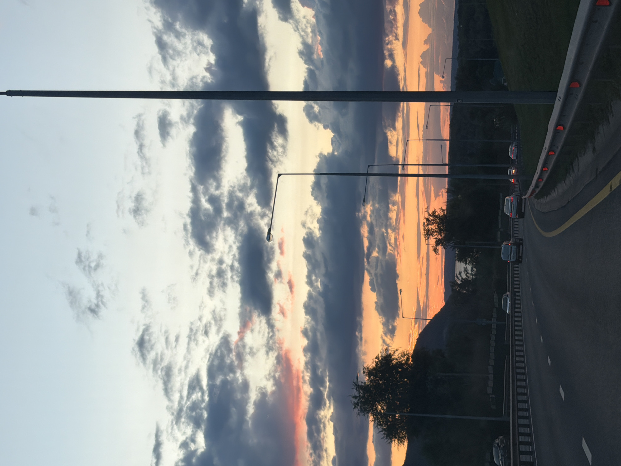 This is a full shot, eye-level, outdoor, vertical photograph of a highway at dusk. The sky is a mix of pale blue and dark gray clouds, with a vibrant orange and pink sunset visible in the distance. A very tall, slender gray pole is on the right side of the frame. Several streetlights are visible along the highway, casting a dim glow. The highway itself is asphalt with white dashed lines indicating lanes. There are guardrails on the right side of the road with red reflectors. Several cars are driving on the highway. In the background, there are dark green trees and hills. The image appears to be taken in a suburban or rural area, possibly outside of Samara, Russia, given the architectural style of the streetlights and the general landscape. There are no people visible in the image.