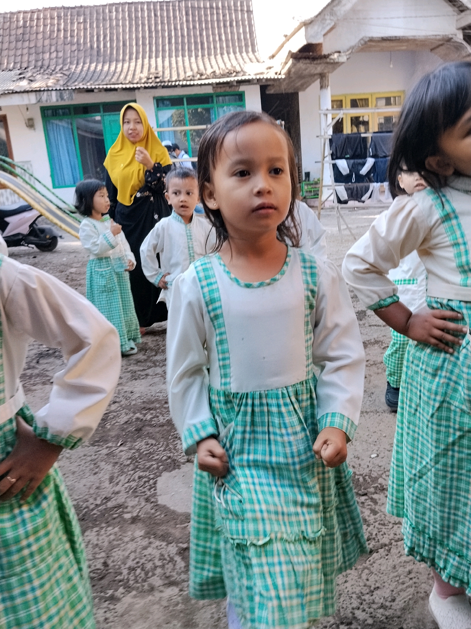 In Panggungan, Indonesia, a group of children, likely students, are gathered outdoors. The central focus is a young girl in the foreground, wearing a white top with a green plaid yoke and a matching green plaid skirt with ruffles. Her expression is neutral, and her gaze is directed slightly off-camera. To her right, another girl, also in a similar green plaid dress, stands with her hand on her hip. In the background, several other children in matching white and green plaid outfits are visible, some appearing to be waiting or observing. A woman in a yellow hijab and black dress stands further back, interacting with one of the children. The environment appears to be a schoolyard or outdoor area with a building in the background, a tiled roof, and a dirt or gravel ground. A scooter is parked to the left, and a rack with hanging clothes is visible near the building. The scene suggests a candid moment during a school day or event.