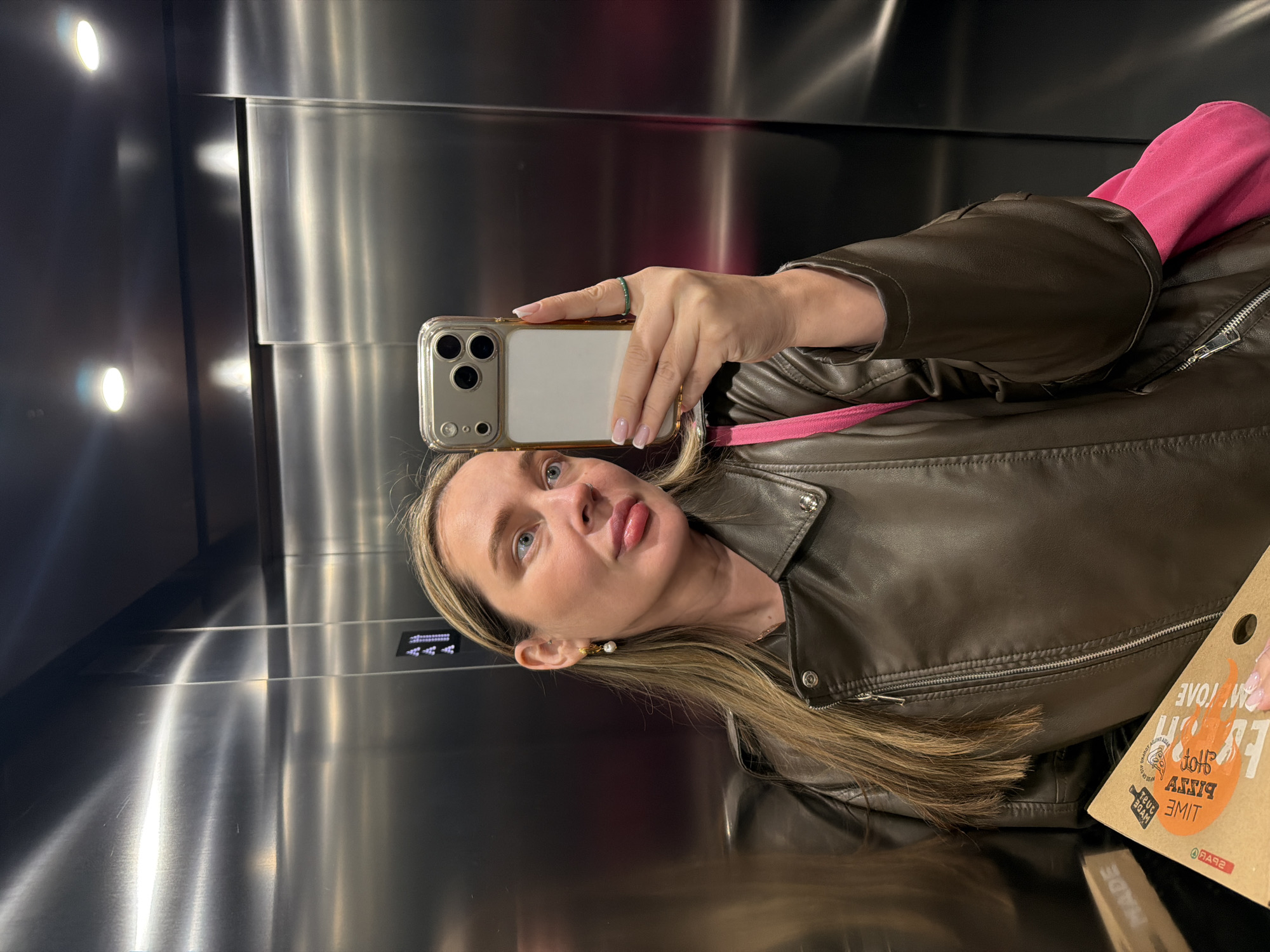 A fair-skinned woman with blonde hair takes a selfie in an elevator. She wears a brown leather jacket over a pink top, a ring on her finger, and a pearl earring. She holds an iPhone in her right hand, partially obscuring her face. Reflected in the background is the metal elevator interior with visible buttons and lights. A pizza box is partially visible on the right side. No specific city or location in Moscow, Russia, can be identified from the available information.