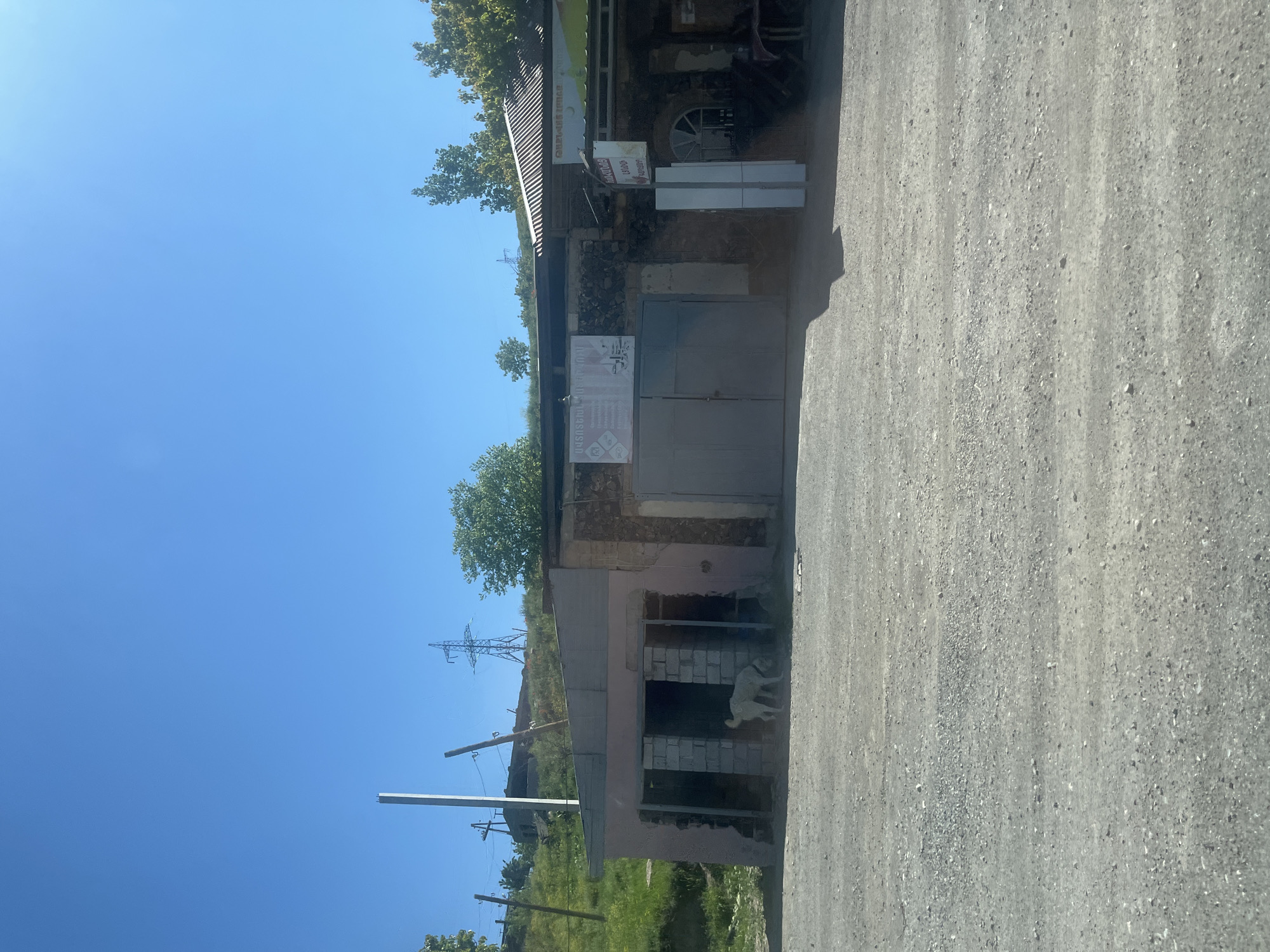 The image captures an outdoor scene under a clear, bright blue sky, indicating daytime. In the foreground, a light gray, gravelly road stretches across the frame. To the left, a single-story building serves as the main subject. It features a corrugated metal roof and walls constructed from a mix of stone, concrete, and plaster, showing signs of wear and possibly incomplete construction or renovation.
The front of the building has large, closed metal gates, likely for a garage or entrance. Above these gates, a prominent white sign displays text in Armenian script: "ԿԱՀՈՒՅՔԻ ԱՐՏԱԴՐԱՄԱՍ," which translates to "Furniture Workshop." A smaller white sign to its right also contains Armenian text, possibly including "ԴՌՆԵՐ" (Doors). Along the side of the building, an open section reveals an interior built with cinder blocks, where a light-colored dog stands, looking outward. Another partially visible sign on this side also features Armenian script and a circular logo.
The setting appears to be a rural or semi-urban area. Beyond the building, green, tree-covered hills rise in the background. A large electricity pylon (lattice tower) is visible on a distant hillside, and several wooden utility poles with wires run closer to the building, indicating utility infrastructure. The scene is static, with no overt activity taking place other than the dog observing its surroundings. The bright sunlight suggests a warm, clear day. The Armenian text on the signs indicates the location is likely in Armenia or an Armenian-speaking region.