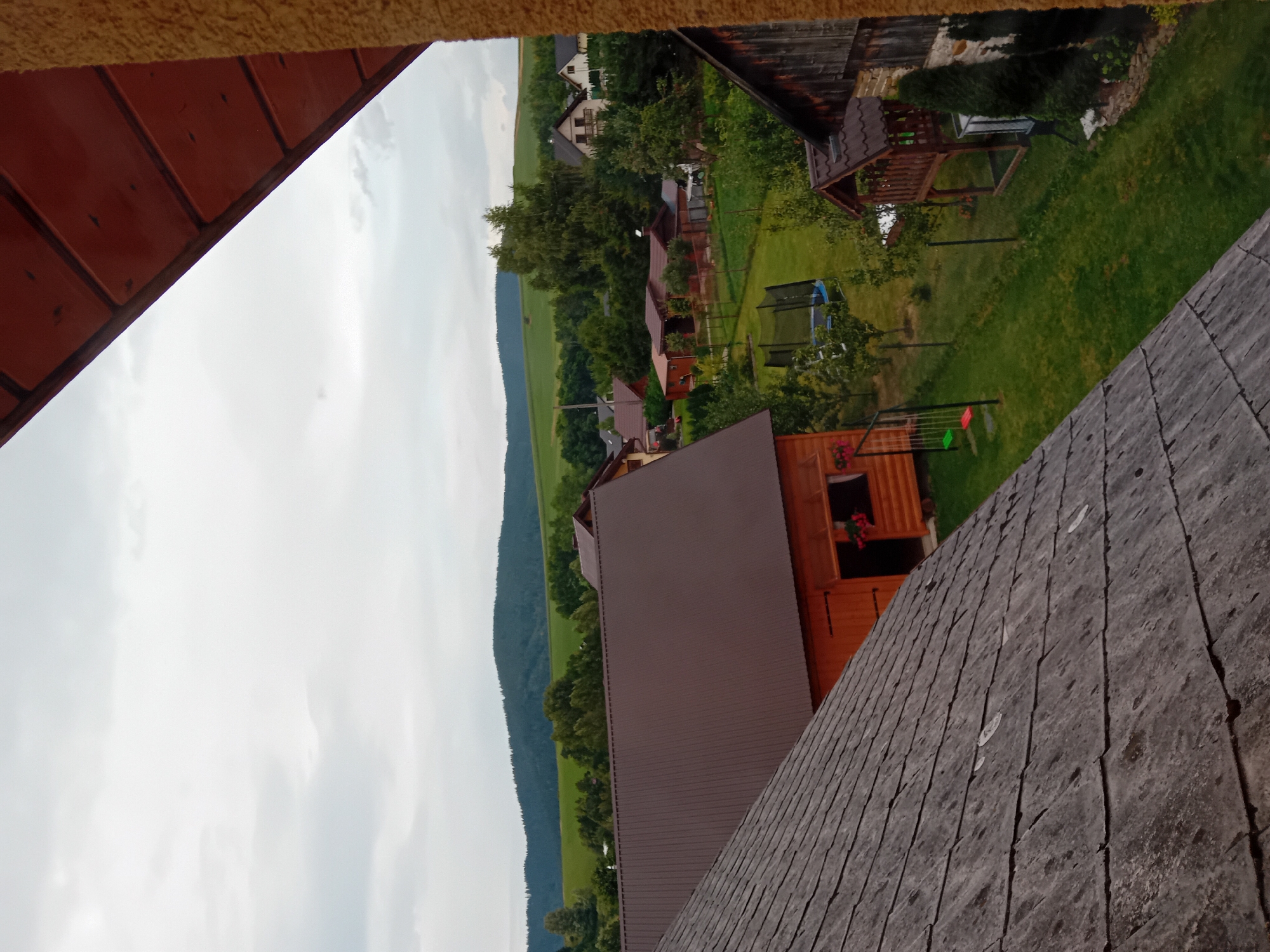 The image shows a panoramic view of a rural village, likely in Zuberec, Slovakia, with several houses, lush green fields, and rolling hills in the background. The foreground features the roof of a house with weathered, gray shingles, partially obscuring a view of a neighboring wooden house with a dark brown metal roof. A small wooden shed with a window is visible on the side of the wooden house. In the middle ground, a well-maintained lawn is dotted with trees and a trampoline. Several other houses with different architectural styles are scattered throughout the scene. The sky is overcast with gray clouds.