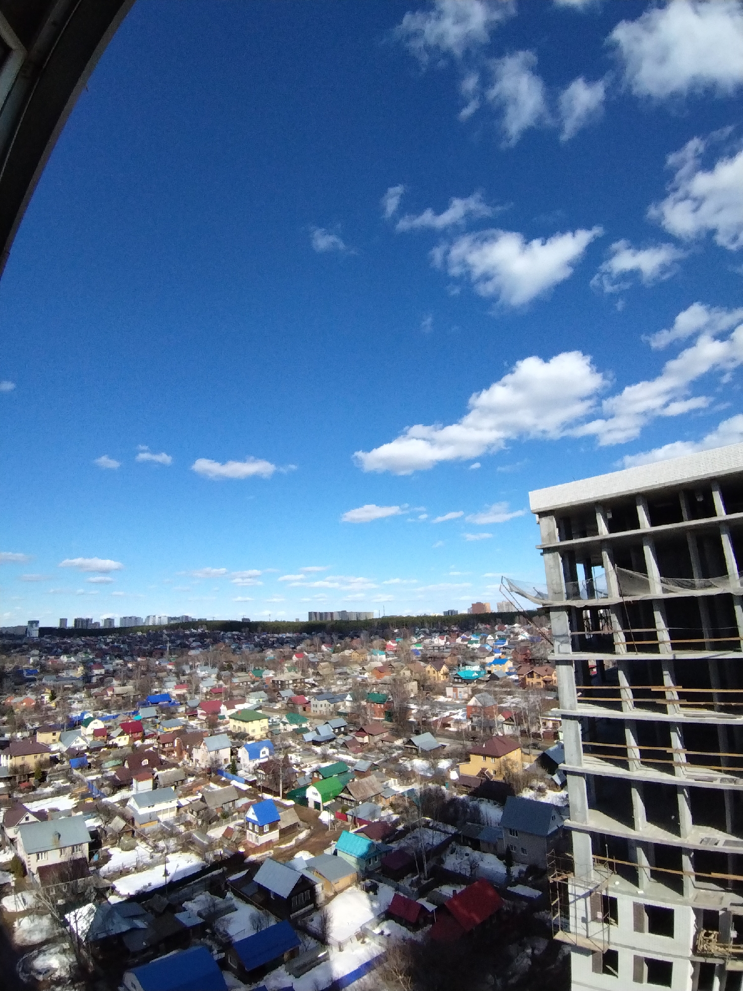 The image presents a view from a high vantage point, possibly from within a building. The foreground features a concrete structure under construction, with its skeletal framework prominently visible. Beyond the construction site, a sprawling residential area with numerous houses, distinguished by their colorful roofs of red, blue, green, and brown, is spread out. Patches of snow dot the ground, indicating a late winter or early spring setting. In the distance, the cityscape extends, blending into the horizon under a bright blue sky with scattered white clouds. The scene appears to be in Izhevsk, Russia, as suggested by architectural styles and the urban layout visible.