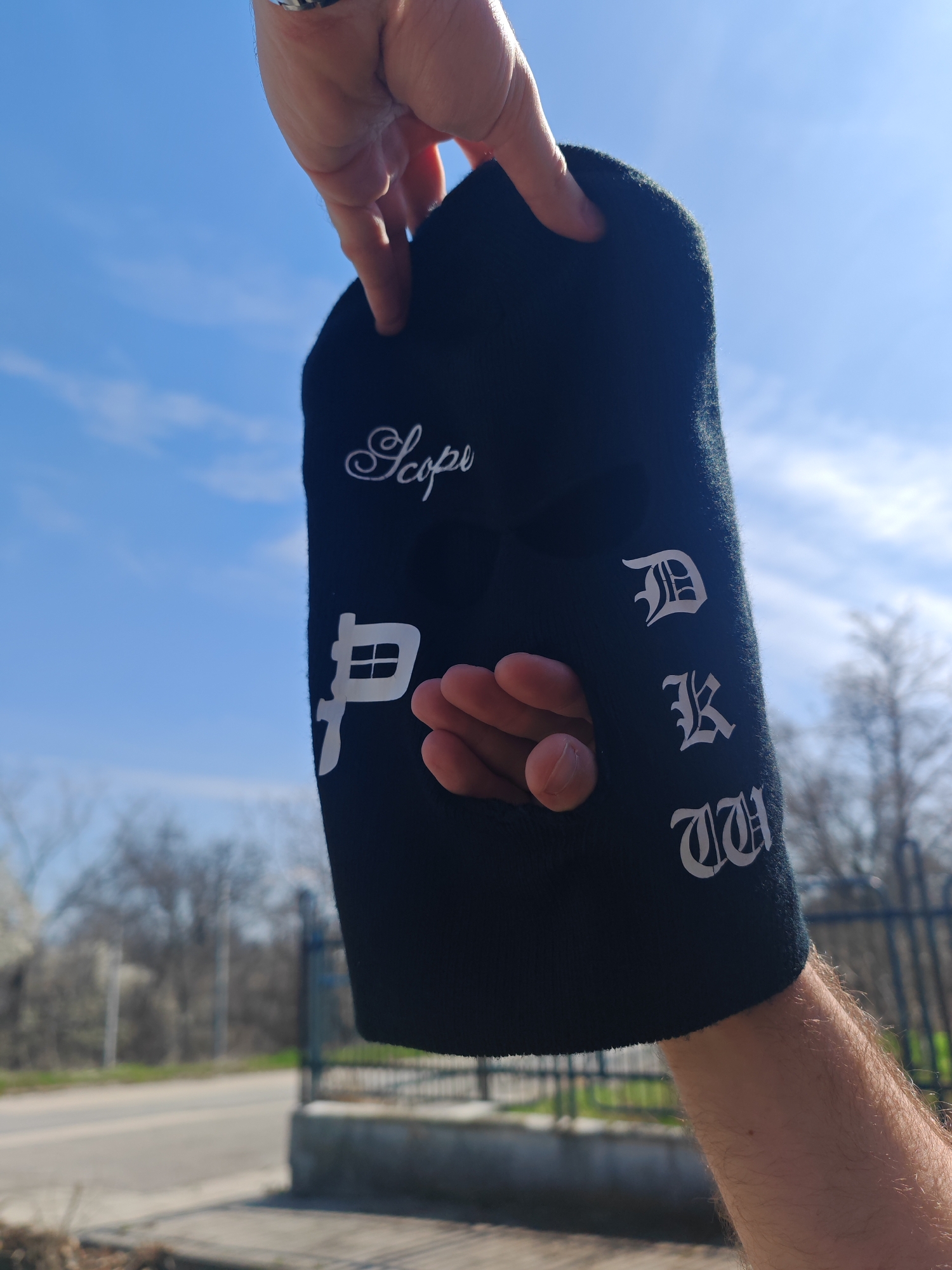 A person's hand is prominently displayed, holding a black balaclava against a bright blue sky with sparse clouds. The balaclava, made of a dark, possibly knit material, is grasped by its top edge between the thumb and index finger, with a silver-colored ring visible on one of the fingers. Another finger, likely the ring finger, from the same hand is inserted into the mouth opening of the balaclava. The person's forearm, fair-skinned and covered with light hair, extends downwards from beneath the balaclava. The balaclava features three openings: two eye slits (one partially visible at the top left) and a mouth opening. White text is embroidered or printed on the balaclava: 'Scope' in a cursive script at the upper left, a stylized character resembling a 'P' with an internal grid structure on the mid-left, and the letters 'D', 'K', 'W' vertically aligned in a Fraktur/Gothic script on the right side. The background, out of focus, reveals an asphalt road or path, a metal fence with vertical bars, and deciduous trees, some with emerging green foliage, suggesting an outdoor setting during late winter or early spring. This scene is located in the city of Shumen, Bulgaria.