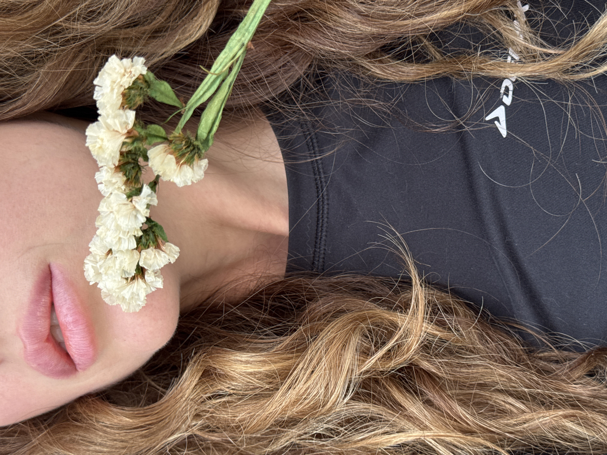 A close-up, oblique-angle shot features a person's face partially obscured by a spray of small white flowers with green stems, resting near their lips. The person is lying down, surrounded by voluminous, light-brown hair with golden highlights. The upper torso is visible, clothed in a dark, likely black, athletic top with a white logo. The location, Chaikovsky, Russia, is irrelevant to the visual content of the image.