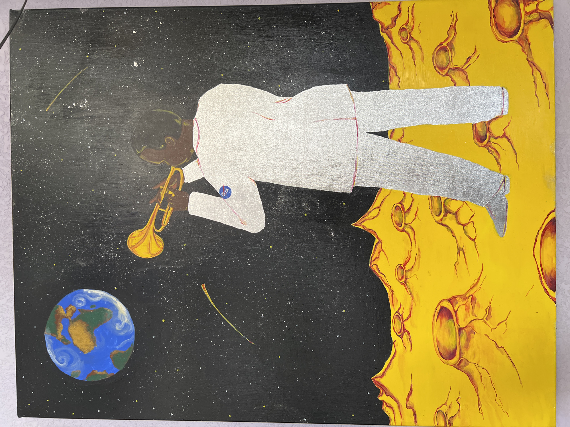 An acrylic painting depicts a celestial scene. A dark-skinned individual stands centrally on a yellow, cratered lunar-like landscape, facing away and slightly turned to the right. The individual wears a shimmering silver suit detailed with faint red piping, bearing a circular blue and white NASA emblem on the left shoulder. Both hands hold a golden trumpet to the individual's mouth, implying the act of playing. The background is a dark, star-filled expanse with numerous white and yellow specks, and two arcing yellow and red streaks. In the upper left quadrant, a stylized blue and green Earth globe is visible. The image is of a physical painting located in Nha Trang, Vietnam.
