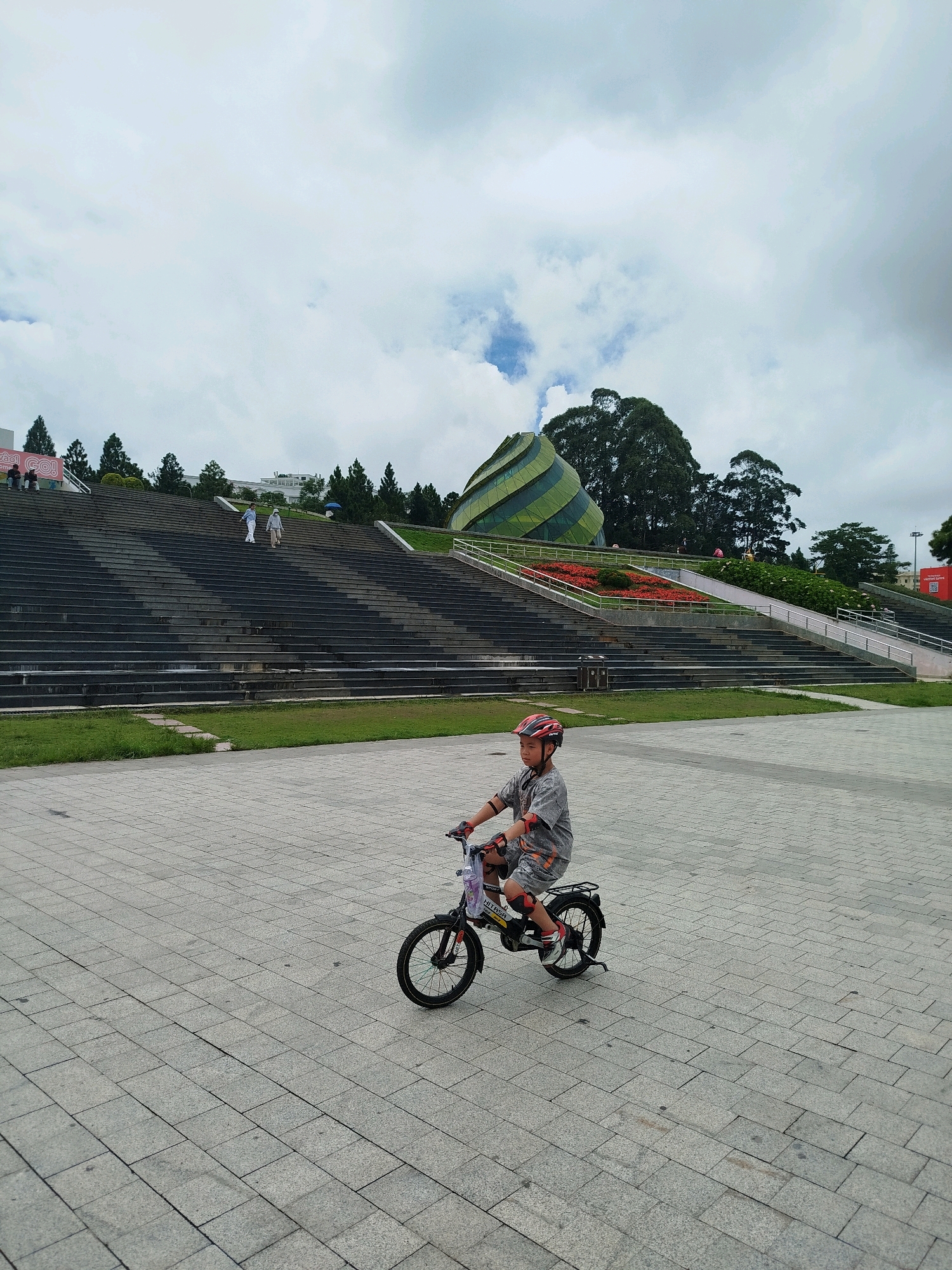 A young boy wearing a grey t-shirt, grey shorts with an orange pattern, a red helmet, and red elbow pads rides a black bicycle on a paved plaza. The boy is positioned in the lower left quadrant of the image, facing right. Behind him, a wide, dark stone staircase stretches upwards. At the top of the stairs, several individuals are walking. Beyond the stairs, a distinctive green and yellow striped, conical building with a curved roof is visible, surrounded by trees and a flowerbed. The sky is overcast with white and grey clouds. The overall scene appears to be the Quang Trung Cultural Park in Da Lat, Vietnam, a public space featuring a large amphitheater, modern architecture, and green spaces.