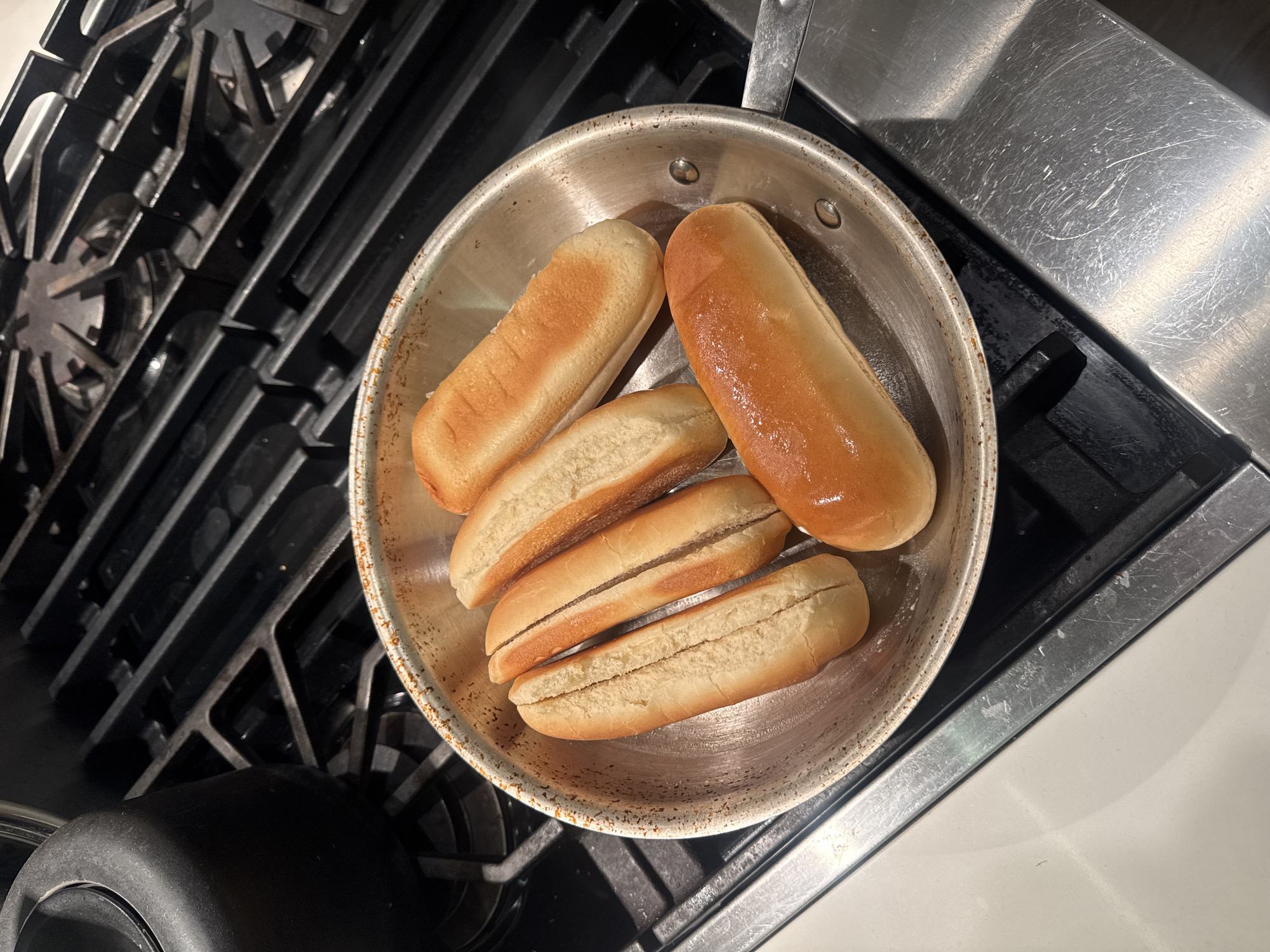 The image shows a stainless steel frying pan containing five hot dog buns, positioned on a stovetop. Two of the buns appear to be whole, while the other three are split open. The stovetop is metallic and shows signs of wear. The setting appears to be a kitchen, possibly in a residential or commercial space in the United States of America.