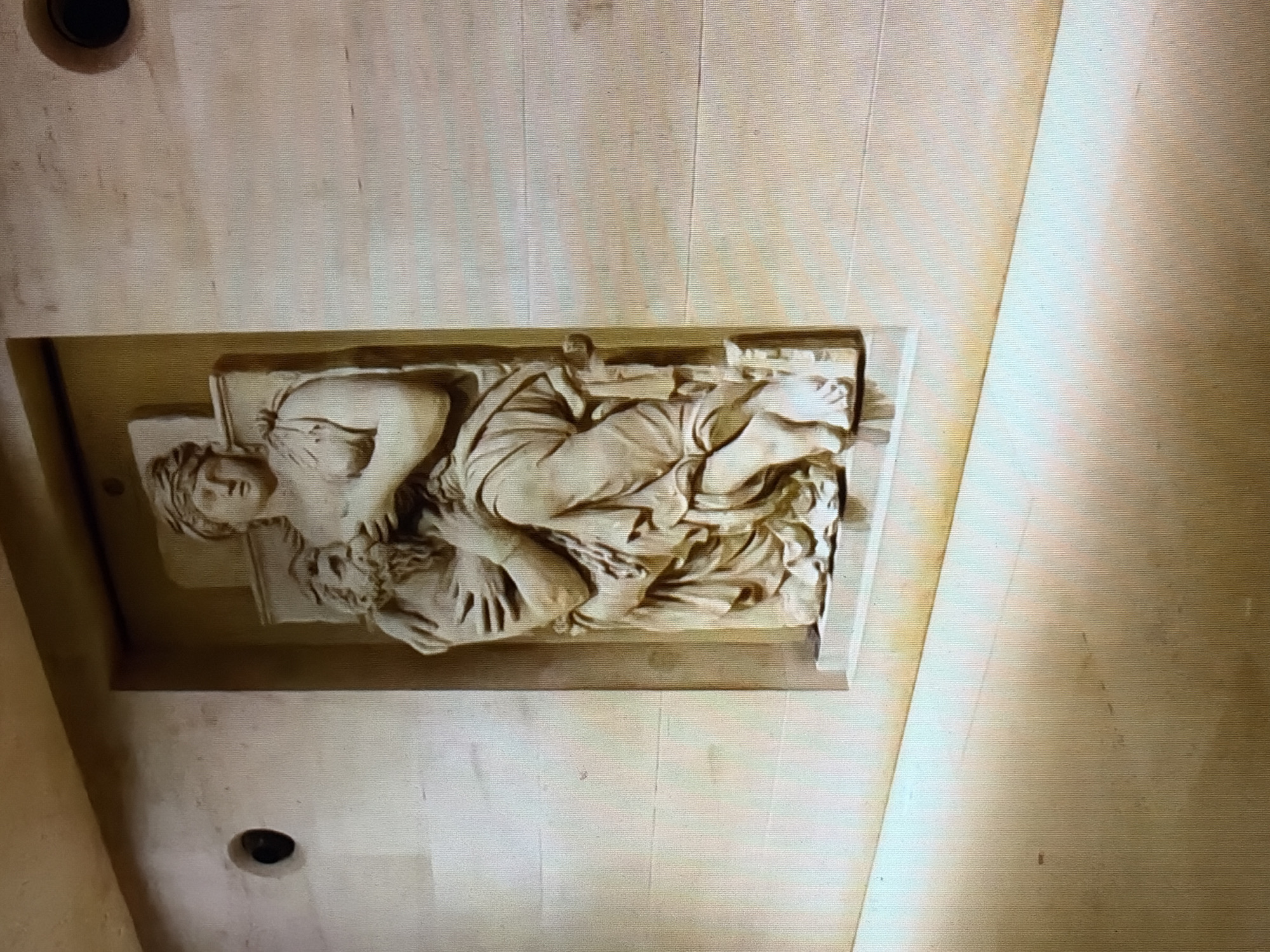 The image shows a low-angle, medium shot of a bas-relief sculpture depicting two figures on a light-colored wall. The sculpture is set within a recessed niche. The figures, carved in a classical style, are a standing woman embracing a seated man. The woman, with a head covering and draped clothing, holds the man close to her chest. The man, who has a beard, appears to be supported by her. Both figures are rendered in stone with detailed musculature and drapery. The wall surrounding the niche is made of large, rectangular stone blocks and has two circular openings or fixtures. The lighting casts shadows, highlighting the three-dimensionality of the relief. The floor in the foreground is also a light-colored, smooth surface, possibly stone or polished concrete, and it slopes downwards away from the wall. The overall environment suggests an interior space, possibly a museum or a historical building.