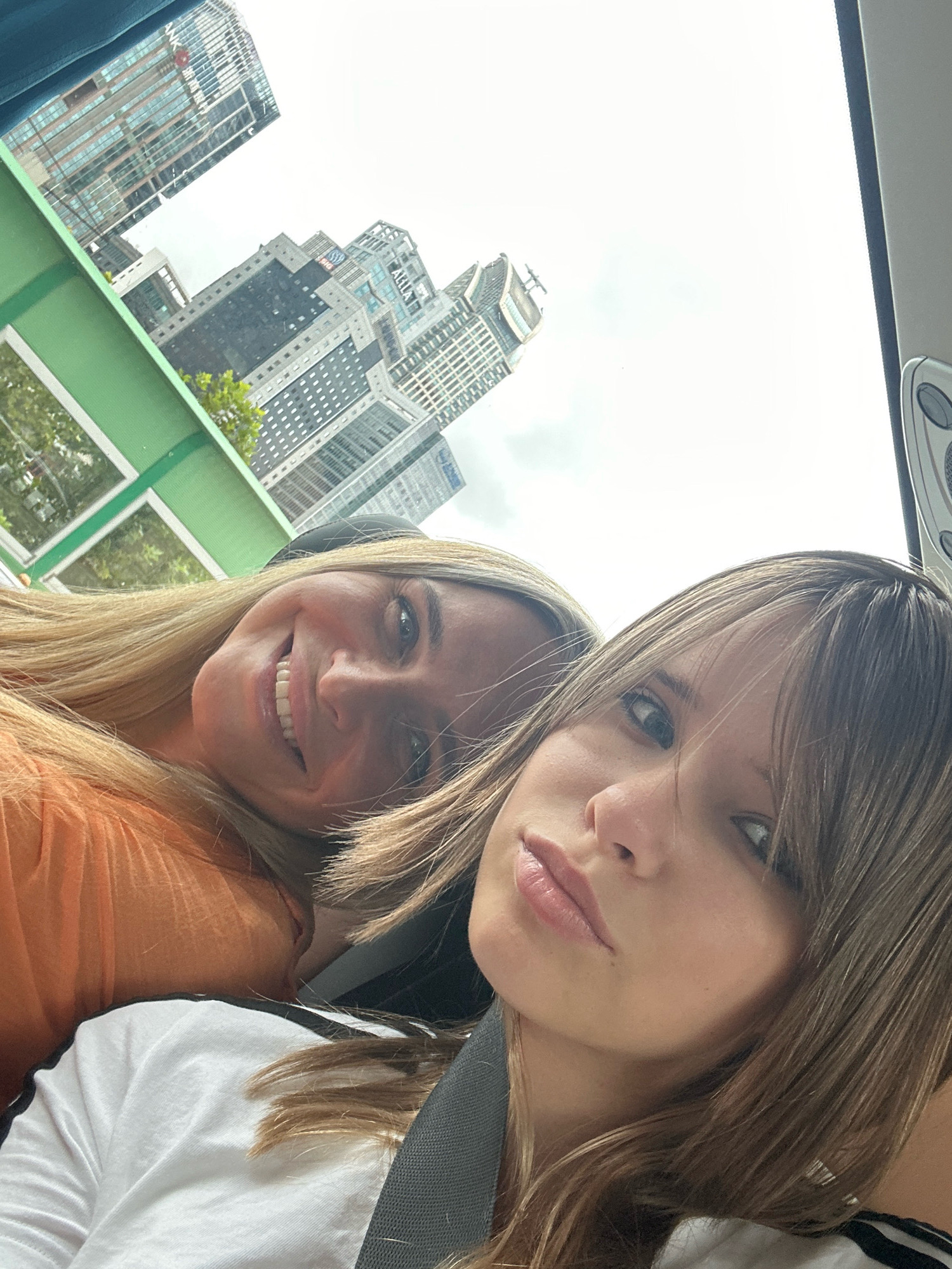 Two individuals, likely a mother and daughter, are captured in a selfie with an urban cityscape in the background. The person in the foreground, on the right, has light brown hair and is looking towards the camera with a slight smirk. They are wearing a white t-shirt with black stripes on the sleeve. The person in the background, on the left, is smiling broadly, with blonde hair, and is wearing an orange top. The background showcases several modern skyscrapers and buildings, suggestive of a dense metropolitan area. The green railing of what appears to be a bus or tram can be seen in the lower left, and the sky is overcast. The scene suggests they are traveling together, possibly on a tour, given the elevated perspective and the glimpse of vehicle interior. The architecture and density of the buildings are characteristic of a major commercial district like Jing'an District, China.