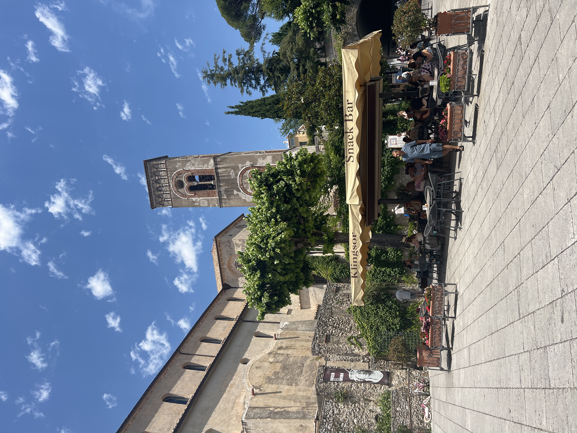 In the city of Ravello, Italy, under a clear blue sky with scattered white clouds, a piazza is bustling with activity on a sunny day. A stone building with arched windows lines the left side of the frame, its weathered facade a testament to history. In front of it, a stone wall is adorned with ivy and a banner depicting a classical bust and Italian text, possibly an advertisement for "Il Museo del Duomo." To the right of the building, a tall, ornate bell tower rises, its architectural details visible against the sky.
In the foreground, a "Snack Bar" is set up under a beige awning, offering a shaded respite for patrons. Numerous tables and chairs are occupied by people enjoying refreshments and conversation. A man in a blue shirt and shorts walks past the cafe, adding to the lively atmosphere. Flower planters with red and pink blooms are strategically placed on the tiled piazza, adding splashes of color to the scene. Bicycles are parked near the stone wall on the left, suggesting visitors have arrived on two wheels. Lush green trees and foliage surround the cafe and the historical structures, creating a picturesque Italian setting.