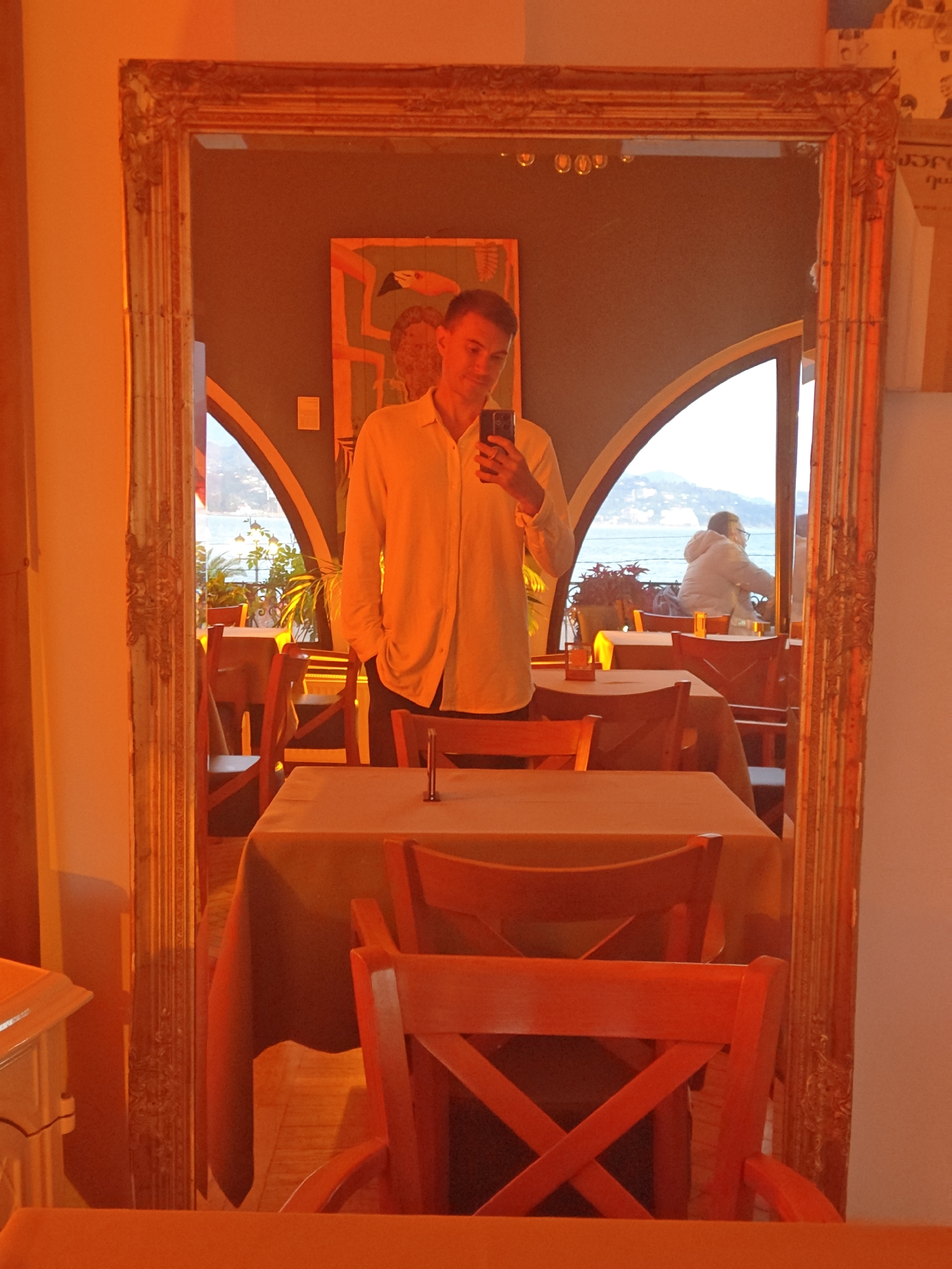 The image captures a mirror selfie taken by a man in what appears to be a restaurant or cafe. A large, ornate gold-framed mirror dominates the foreground, reflecting the scene within the establishment.
The photographer, a man wearing a light-colored, long-sleeved shirt and dark trousers, is centered in the mirror's reflection. He holds a black smartphone up to take the picture, with a neutral expression. The reflection shows an interior space furnished with reddish-brown wooden chairs featuring X-shaped backs and tables draped with light-colored tablecloths. On the wall behind the man's reflection, an abstract or stylized painting, featuring a bird-like figure, is visible.
Through large arched windows or openings in the background of the reflection, a wide body of water, likely the sea or a large lake, is visible, with distant landforms or mountains on the horizon. Another person, viewed from behind, sits at a table near one of these windows, wearing a light-colored puffy jacket and glasses, seemingly enjoying the view. Potted plants are also arranged near the windows.
The interior lighting has a distinct warm, orange, and reddish hue, especially pronounced on the left side of the mirror and its frame, possibly due to ambient lighting or an image filter. The natural light filtering in from the windows appears softer, suggesting a late afternoon or bright, diffused daylight. The setting evokes a serene atmosphere in a scenic waterfront location. Some indistinct text can be vaguely seen on a wall hanging to the far right within the reflection.