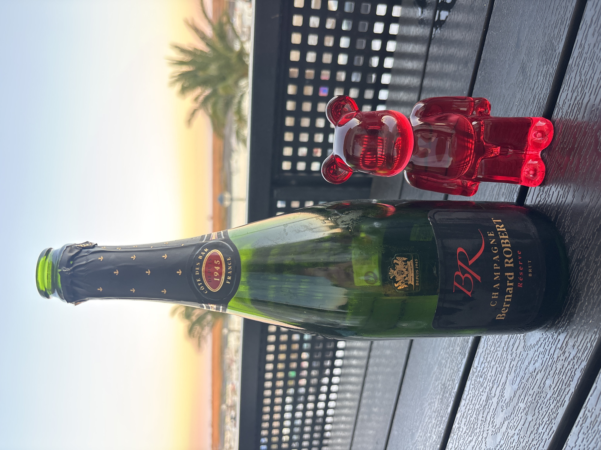 A champagne bottle and a red figurine are placed on a dark wooden table. The bottle is green glass and has a black label with gold lettering that reads "BR CHAMPAGNE Bernard ROBERT Réserve BRUT". The red figurine is a bear-shaped toy with a translucent finish. In the background, there is a blurred view of palm trees, a body of water, and a fence. The sky is clear and shows a gradient of colors, suggesting it is either sunrise or sunset. The scene appears to be an outdoor setting, possibly a restaurant or balcony overlooking a waterfront. There is no indication that this scene is in Tavira, Portugal.