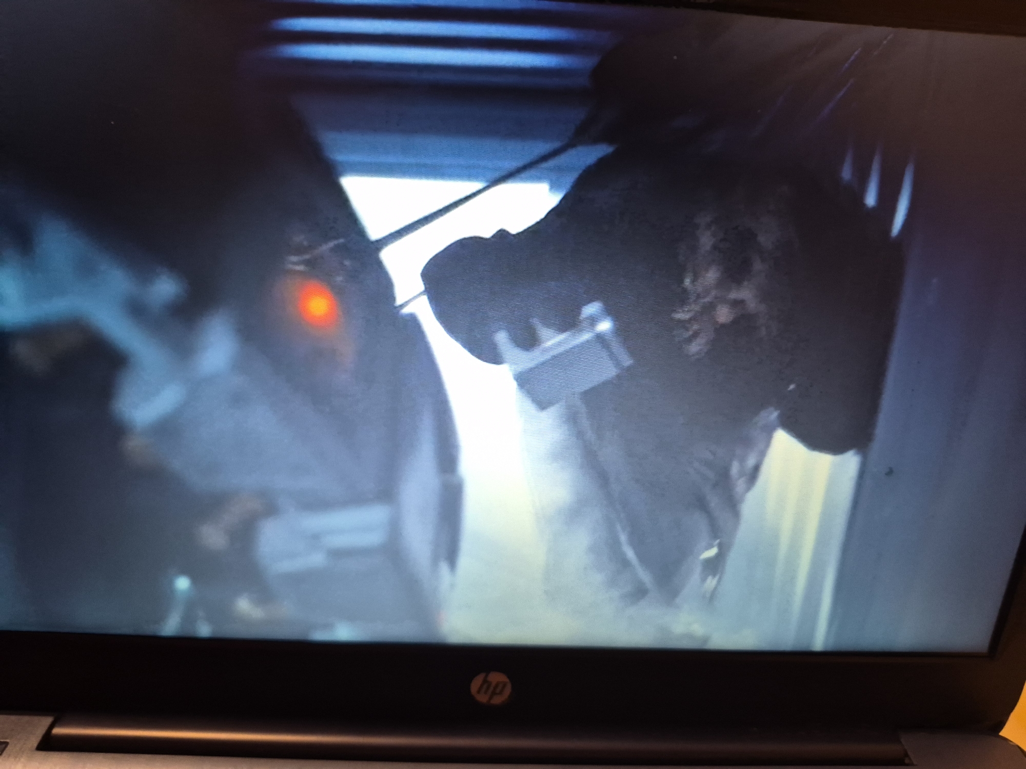 This is a close-up, low-angle, slightly blurry photograph of a laptop screen displaying a scene that appears to be from a video game or movie. In the foreground, the black bezel of the laptop is visible. The screen displays a dark, gritty, and somewhat futuristic environment. On the left side of the screen, a figure with a glowing red eye is visible, suggesting it might be a robot or an alien creature. This figure is partially obscured by shadows and appears to be wearing some sort of armor or clothing. To the right, another figure or object is visible, also in shadow, with some metallic or mechanical elements. The background is a mix of dark and illuminated areas, with what looks like metallic structures or a confined space. There are no identifiable landmarks or elements that would indicate a specific location within Sandwell, United Kingdom.