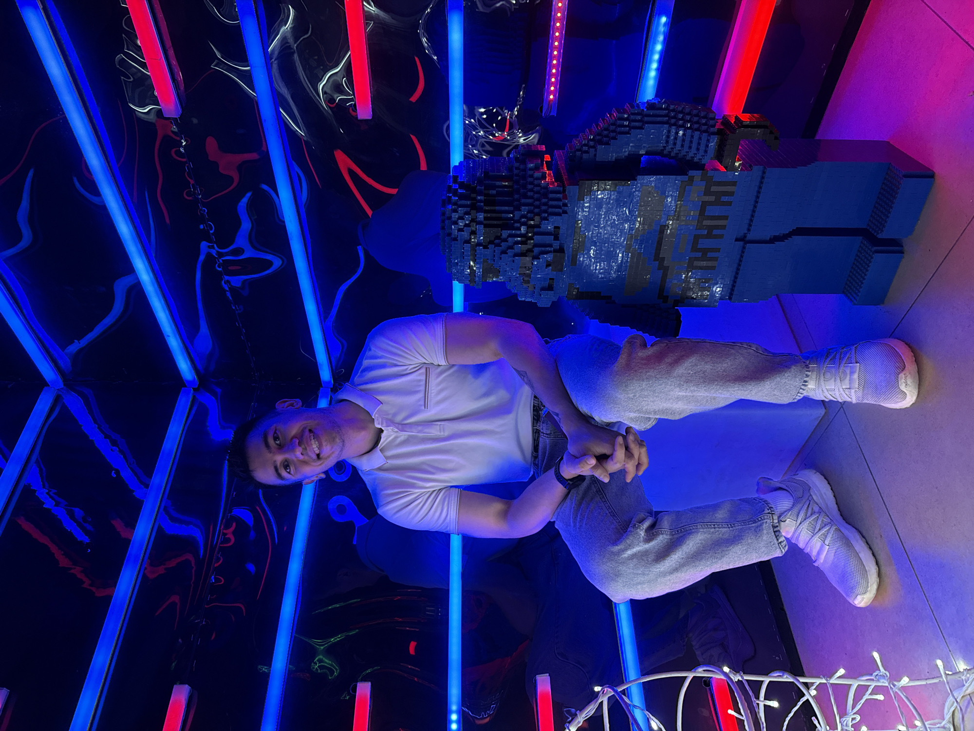 A medium-shot, slightly angled photo shows a man in a white polo shirt and jeans, positioned beside a large Lego sculpture in an indoor space with blue and red neon lights. The man is smiling, his hands clasped in front of him. He is wearing white sneakers. The background is a reflective surface, distorted by the neon lights, with a string of white decorative lights at the bottom. The location is Kazan, Russia.