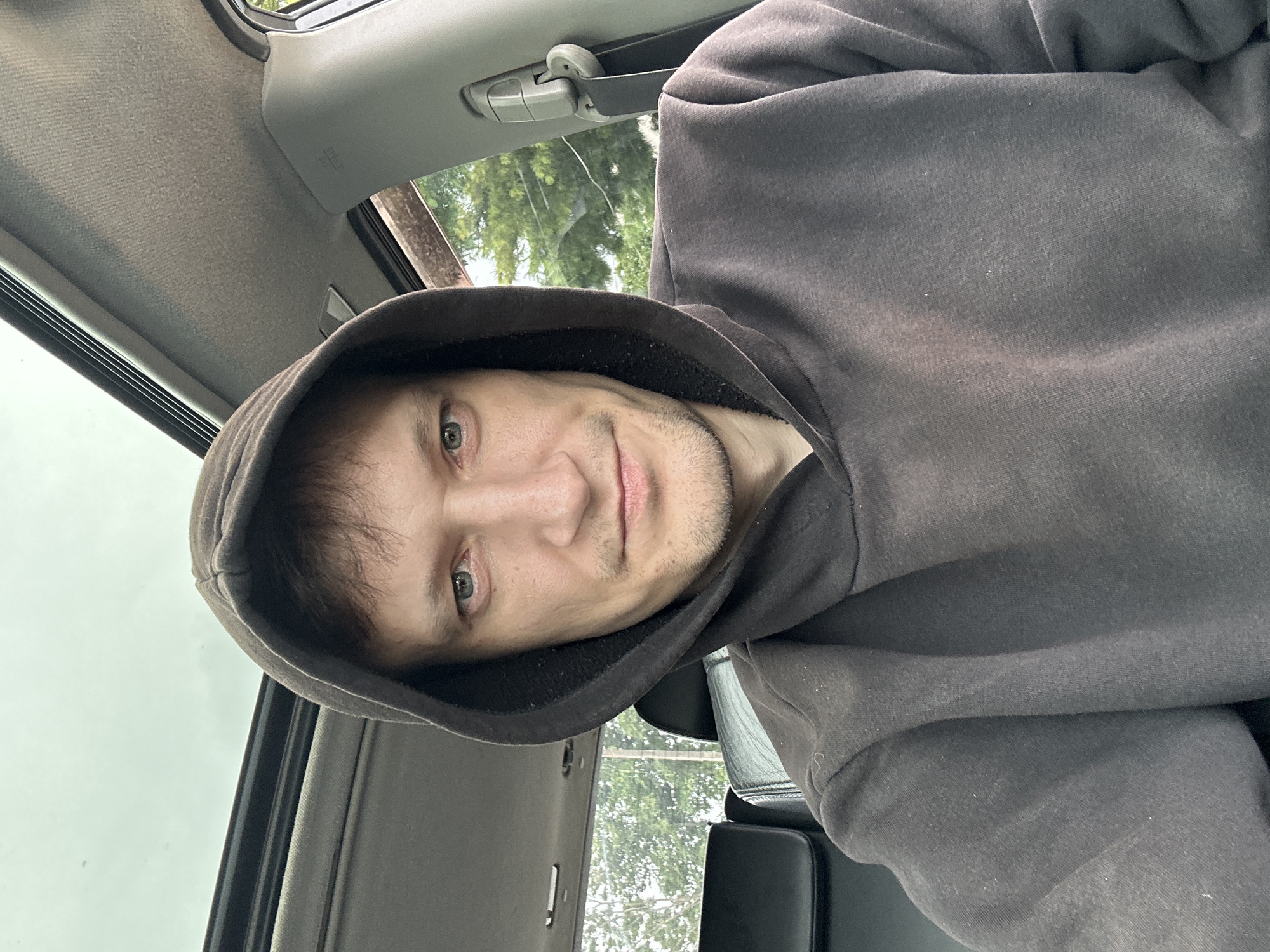 A man wearing a dark hooded sweatshirt is taking a selfie inside a car. The man's face is visible, and he has light-colored eyes and short brown hair. He is looking directly at the camera with a neutral expression. The car's interior is visible, including the sunroof, dashboard, and part of the passenger seat. Through the car windows, trees and foliage can be seen, suggesting an outdoor setting. The image appears to be taken during the daytime. The location is identified as Makiv, Ukraine.