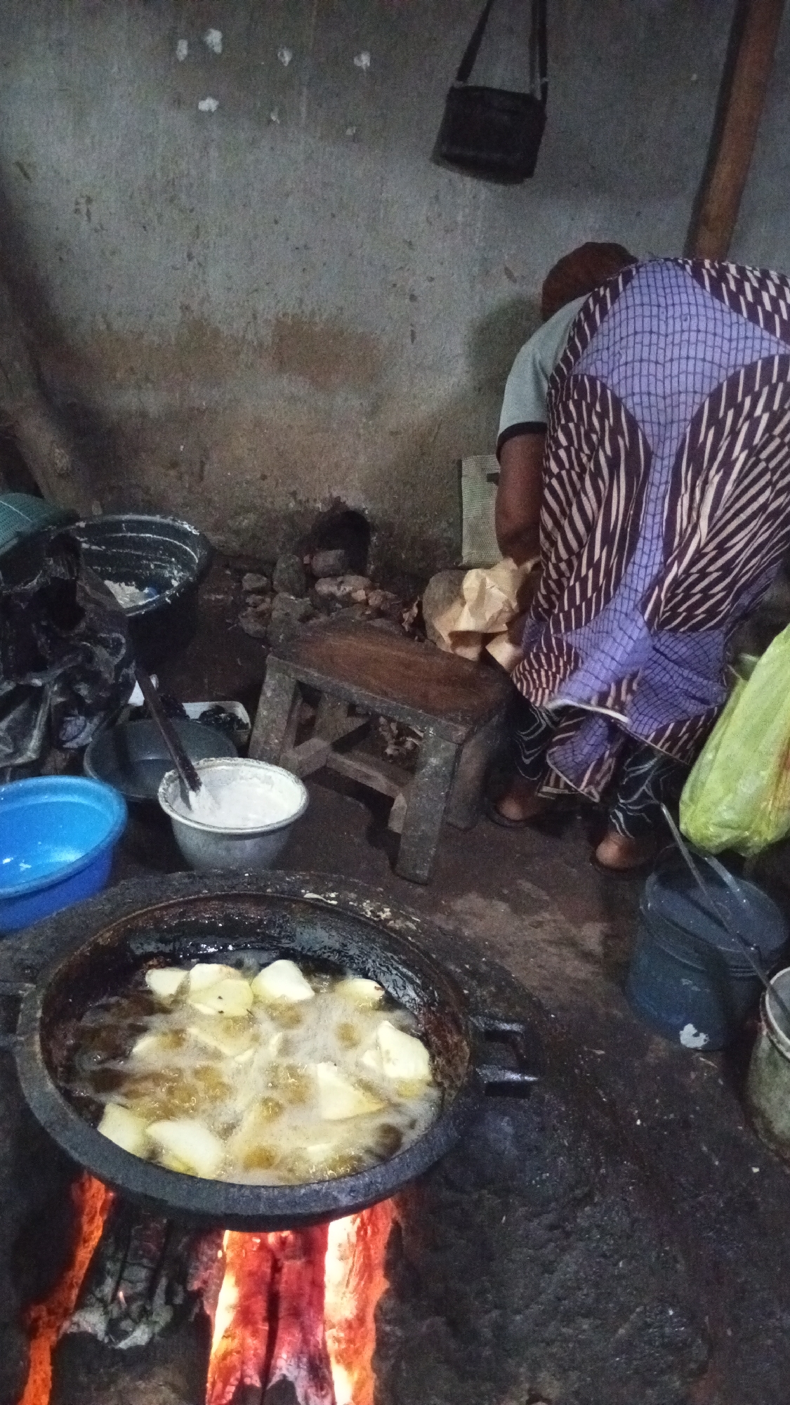 In Adankolo, Nigeria, the image shows a person in a patterned purple skirt standing near a rough wooden stool, potentially preparing food. A black pan sits over an open fire with food frying in oil. A blue bucket and a bowl containing a white mixture are nearby. A handbag hangs on the wall behind the person. The environment appears to be a rudimentary outdoor kitchen or cooking area.