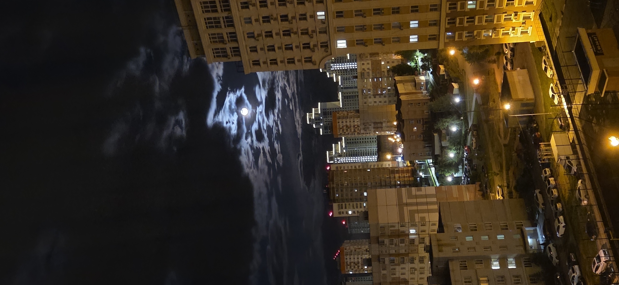 The image shows a nighttime urban scene in an unknown city. The view is from an elevated position, possibly from a high-rise building, capturing the urban landscape below. In the left portion of the image, a dark, cloudy sky is visible with the full moon shining brightly through the clouds. On the right, an array of buildings of various heights dominate the scene. The buildings are lit up with internal lights, giving a warm, yellow glow to their exteriors. Streets and parking areas are also visible, illuminated by street lamps, with several parked cars. There are no people visible in the image. The overall atmosphere is one of a quiet night in the city.