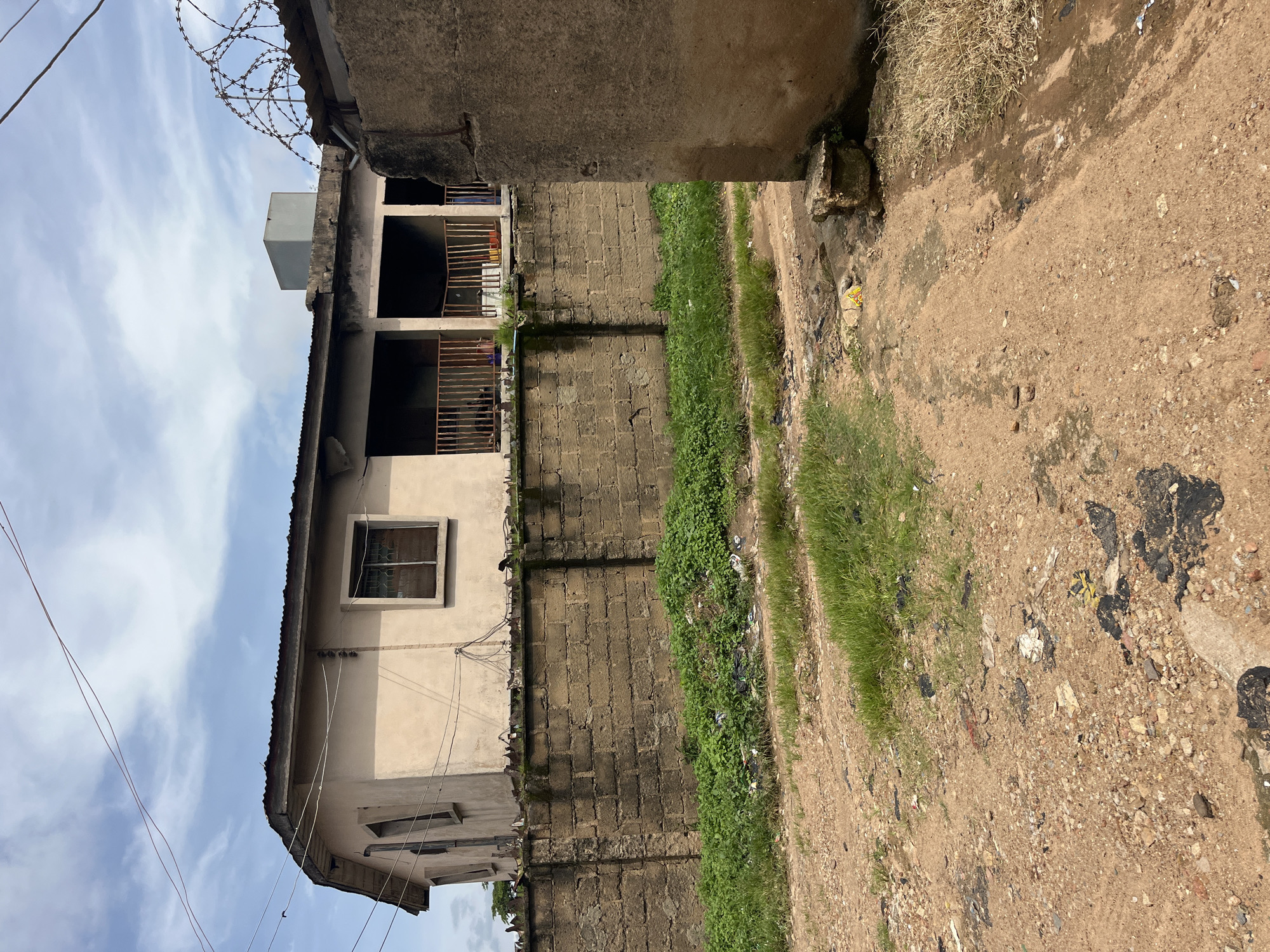 The image depicts an outdoor scene in a somewhat neglected urban environment. A light-colored, two-story building with a corrugated metal roof stands behind a rough, stone-block wall. The building has multiple windows and a covered porch area on the upper level, supported by pillars, with a metal railing. The wall is topped with barbed wire. In front of the wall, there is a strip of overgrown green vegetation mixed with visible litter. The foreground is a dirt and gravel path, also containing scattered debris. A section of a concrete structure, possibly part of another building or wall, is visible on the right edge of the frame. The sky above is partly cloudy. The overall impression is one of a modest, possibly older, residential or commercial area with a lack of recent maintenance. This scene is located in the city of Jos, Nigeria.