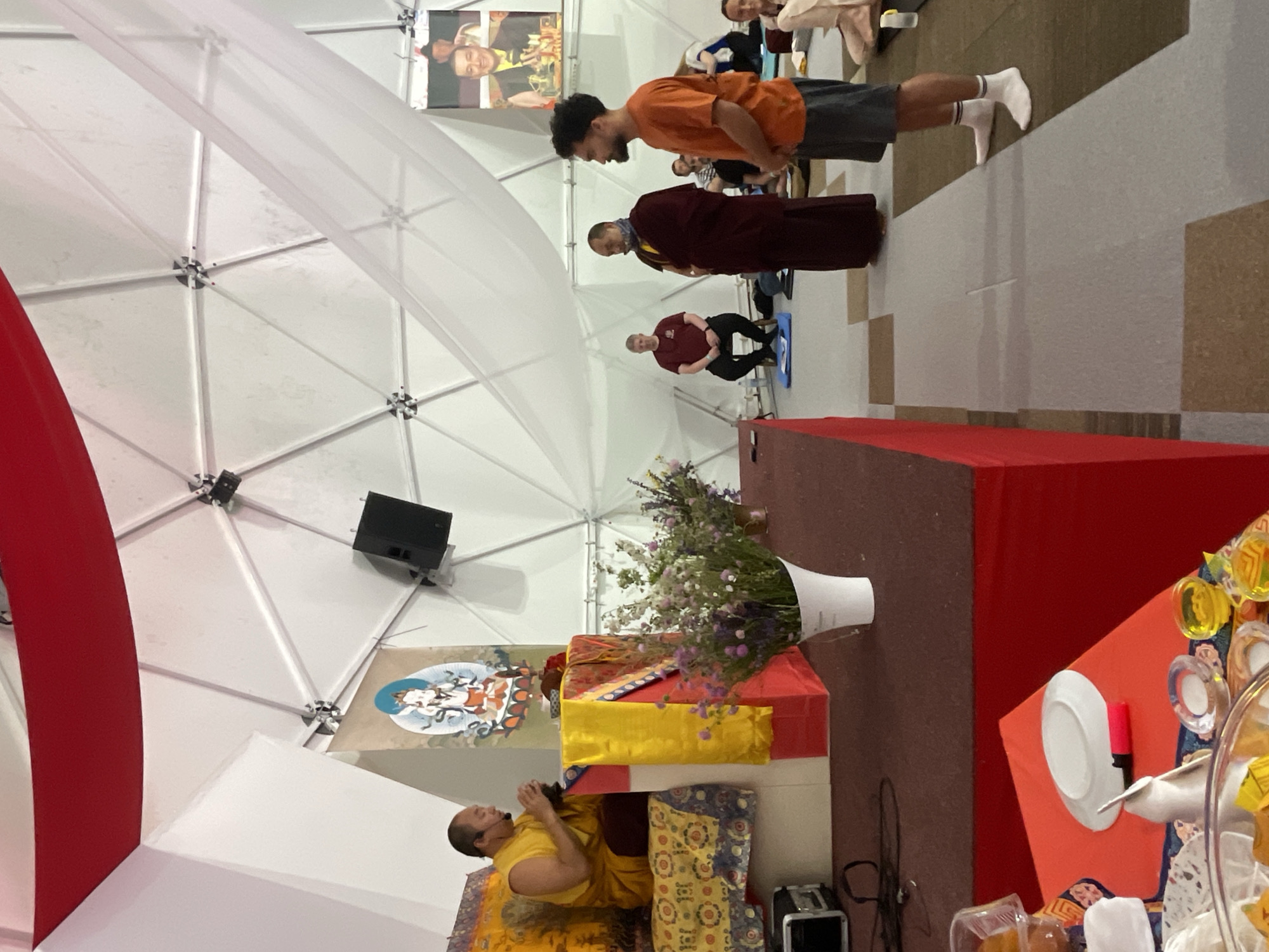 The image depicts an event, possibly a ceremony or performance, taking place indoors under a large white dome-like structure with visible support beams. The location is suggested to be in or around Mekića Kula, Montenegro.
Several individuals are visible. A man in an orange shirt and grey shorts stands with his head bowed, followed by a person in maroon monastic robes, and another individual in a maroon shirt sitting on a blue mat. Further in the background, more people are seated.
In the foreground, a monk in yellow robes is sitting on the floor with his hands in a prayer position, wearing a headset microphone. He is seated next to a red-and-brown-covered raised platform. On the platform is a vase of flowers, some religious objects covered in yellow and orange fabric, and a table with various objects including plates, cups, and other unidentified items. A speaker is attached to the white dome structure above the platform.
Other elements include decorative red fabric hanging from the dome structure and a poster of what appear to be religious figures. The floor is covered with carpet with a repeating pattern.