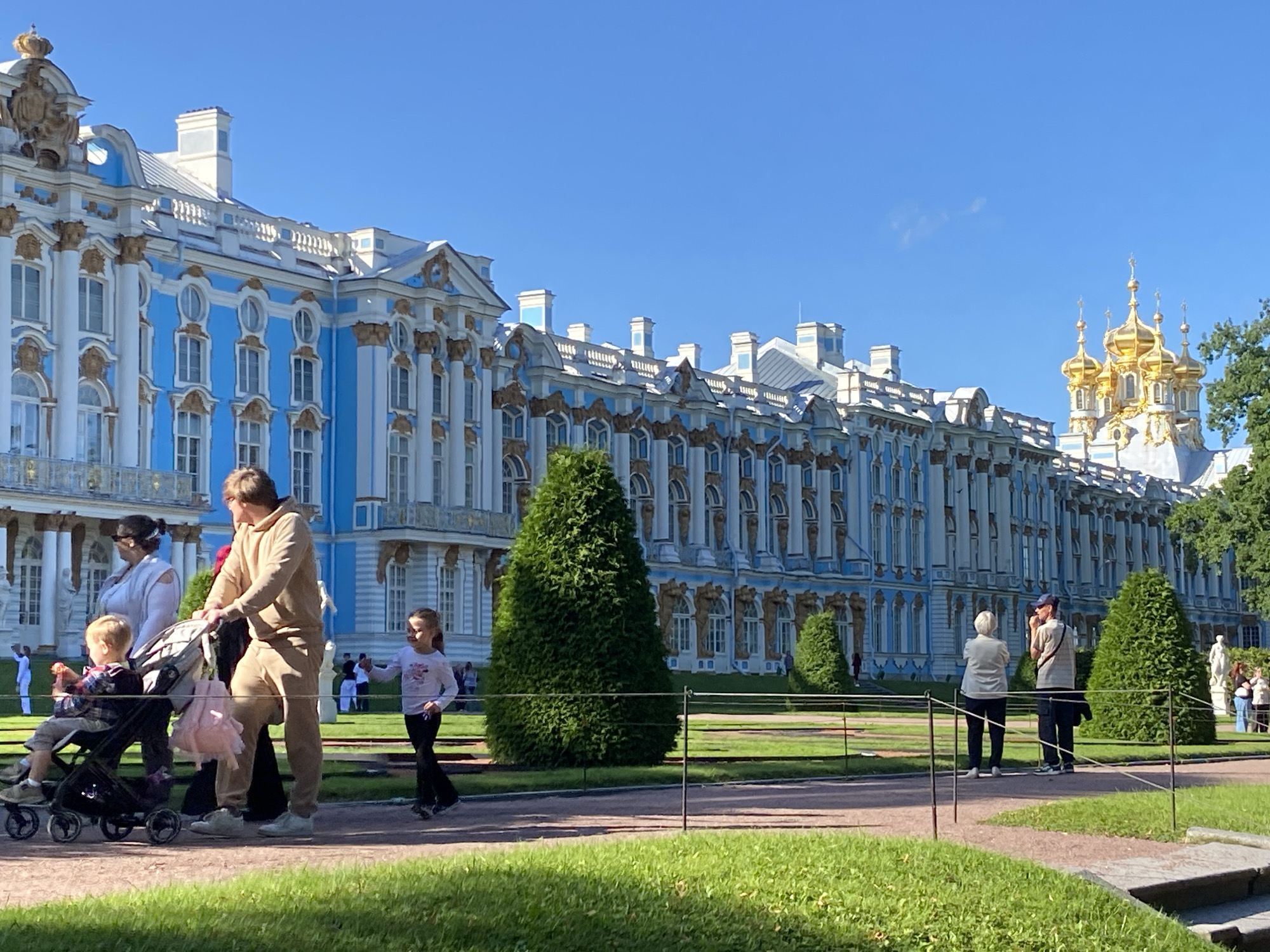 The image depicts the Catherine Palace in Pushkin, Russia, under a clear blue sky. The palace, painted a vibrant blue with white and gold architectural details, stretches across the background. In the foreground, several people are visible. A man and a woman walk with a young child in a stroller, and a young girl runs beside them. Further in the background, a few more individuals are seen walking or standing on the grounds. The palace is surrounded by a manicured lawn and trees, with a pathway in the immediate foreground.