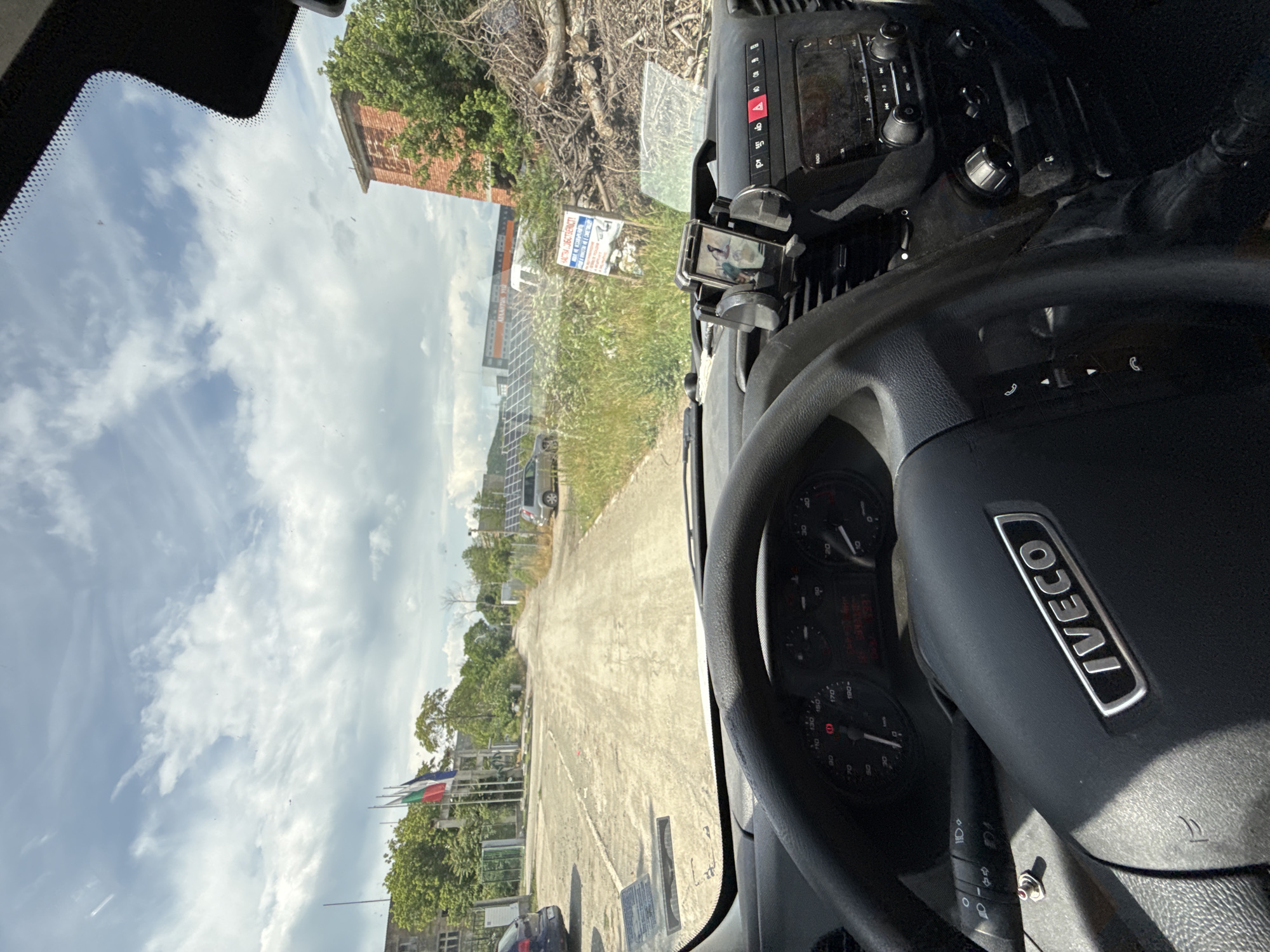 The image captures a driver's perspective from inside an Iveco commercial vehicle, looking out through the windshield on a bright, partly cloudy day. The original image is rotated 90 degrees, so viewing it upright shows the steering wheel on the right, indicating a left-hand drive vehicle.
The interior of the vehicle reveals a black steering wheel with a metallic "IVECO" logo. Behind it, the dashboard instruments, including a speedometer and tachometer, are partially visible. To the right of the steering wheel, the center console features an infotainment system with a screen and various knobs and buttons, including a prominent red hazard light button. A smartphone is mounted in a holder to the right of the console, displaying an image of what appears to be a bird or small animal.
Outside the vehicle, a concrete or dirt path extends into the distance. To the left of the path, a red-brick building is partially seen through trees, and further back, another industrial-looking building. A large array of solar panels is installed along this side. A white sign in Bulgarian is clearly visible, displaying "MUTA - ЖИВОТНОВЪДСТВО" (MUTA - LIVESTOCK FARMING), with additional text such as "ПРОДАЖБА" (SALE) and "МЛЯКО" (MILK).
On the right side of the path, several flagpoles display flags; one has vertical red, white, and green stripes, another is blue, and a third is red, all partially obscured by green foliage. A dark-colored car is parked in the foreground near the flags. The sky is bright with scattered white clouds, suggesting mid-day. The scene depicts a semi-rural or industrial area, consistent with the location context of Kalitinovo, Bulgaria, hinting at an agricultural or farming facility.