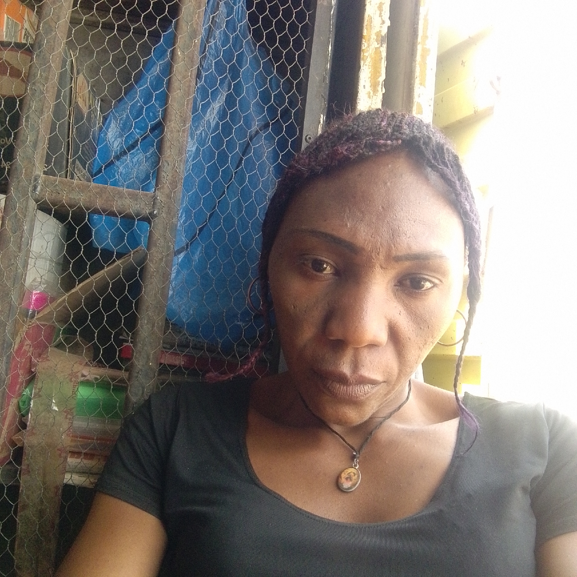 A medium-shot photograph depicts a woman indoors, possibly in the city of Jos, Nigeria. She wears a dark-colored, short-sleeved shirt and a necklace with a pendant. Her hair is braided, with a hint of purple visible in some strands. Behind her, a wire mesh structure is visible, with what appears to be blue plastic sheeting and various objects and containers stacked behind it. The background also contains a partially visible, worn window frame. The lighting is bright, possibly natural light from the window.