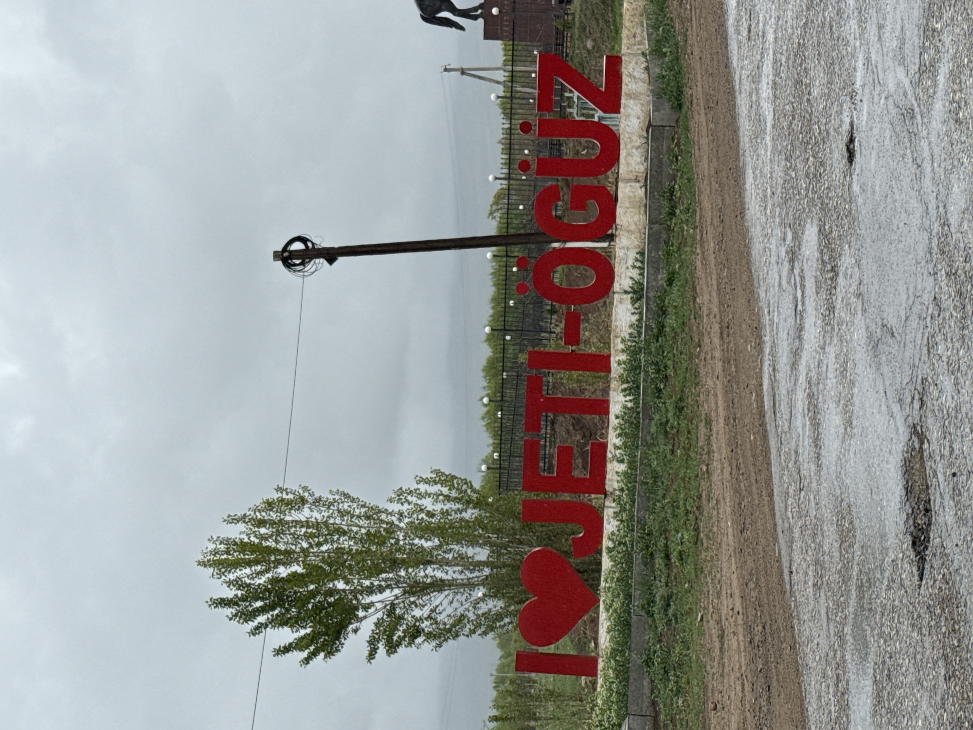 The image depicts an outdoor scene under an overcast sky, likely capturing an entrance or landmark in Jeti Oguz, Kyrgyzstan. The central subject is a large, red, three-dimensional sign spelling out "I ❤️ JETI-ÖGÜZ", oriented vertically from bottom to top in the original image but implying a left-to-right reading when viewed conventionally. The "I" is followed by a prominent red heart symbol, and then the letters "JETI-ÖGÜZ".

The sign is situated alongside a paved road, which appears wet or damp, suggesting recent rain. To the left of the road is a dirt shoulder with green grass and small plants growing, bordered by a concrete curb. Behind the sign and vegetation, a dark metal fence with evenly spaced small, round lights stretches across the mid-ground.

In the left foreground, a deciduous tree with green foliage stands, partially obscuring the "I ❤️" portion of the sign. A wooden utility pole with coiled wires at its top stands between the tree and the "JETI-ÖGÜZ" part of the sign. In the background, beyond the fence, is a distant landscape that appears hazy under the grey, cloudy sky. A dark, partially visible equestrian statue on a pedestal can be seen near the top right, behind the "ÖGÜZ" section of the sign.

The overall atmosphere is subdued due to the cloudy weather, with diffused lighting. No people or active events are visible, presenting a static landscape view of this location.