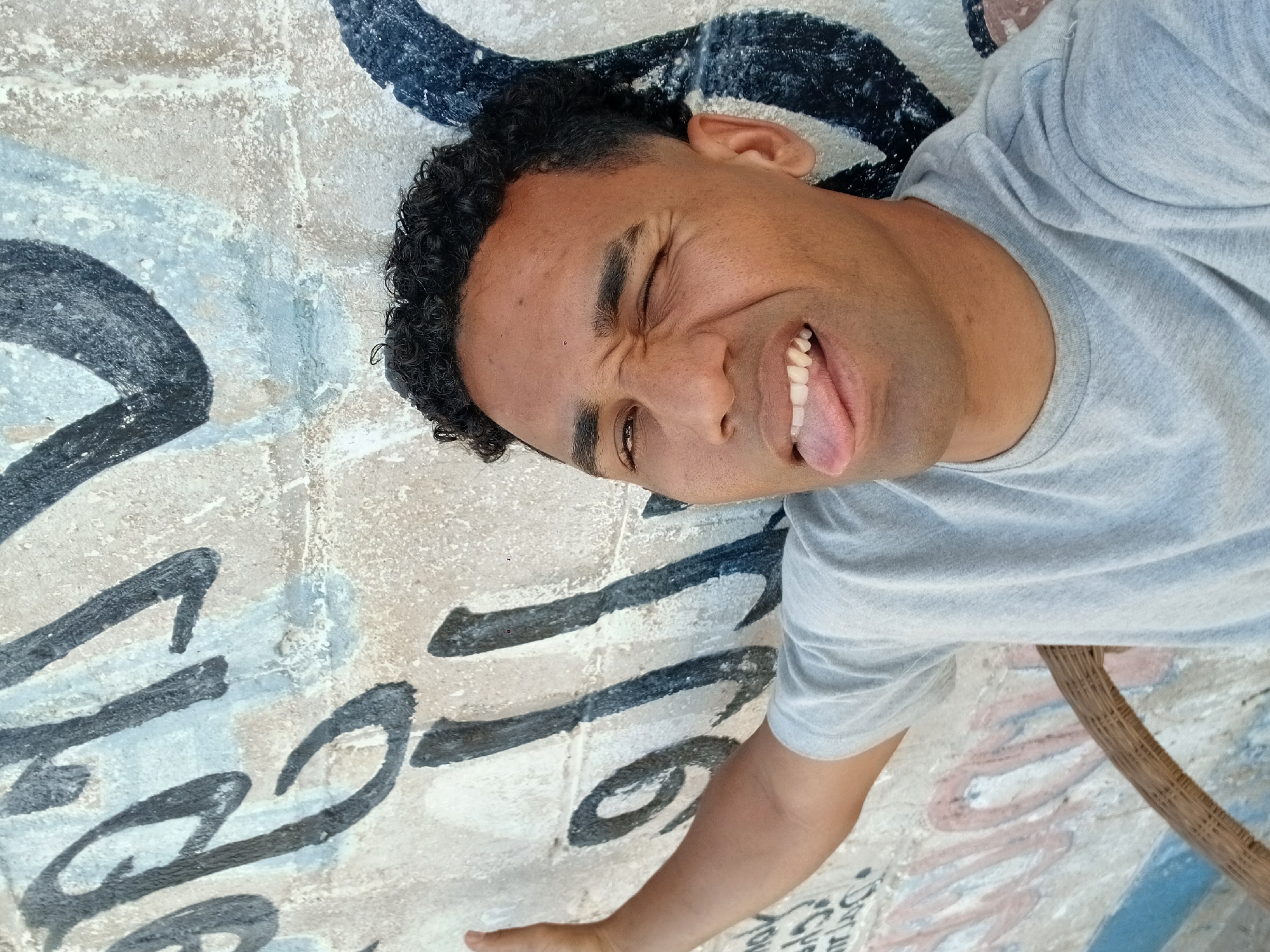 A person with short curly black hair is lying on a graffiti-covered wall in Porlamar, Venezuela. The person is wearing a gray t-shirt and has their tongue sticking out and eyes squinted, making a face. The wall has black and white graffiti markings on a faded background. A wicker-like object is visible in the bottom right corner of the image.