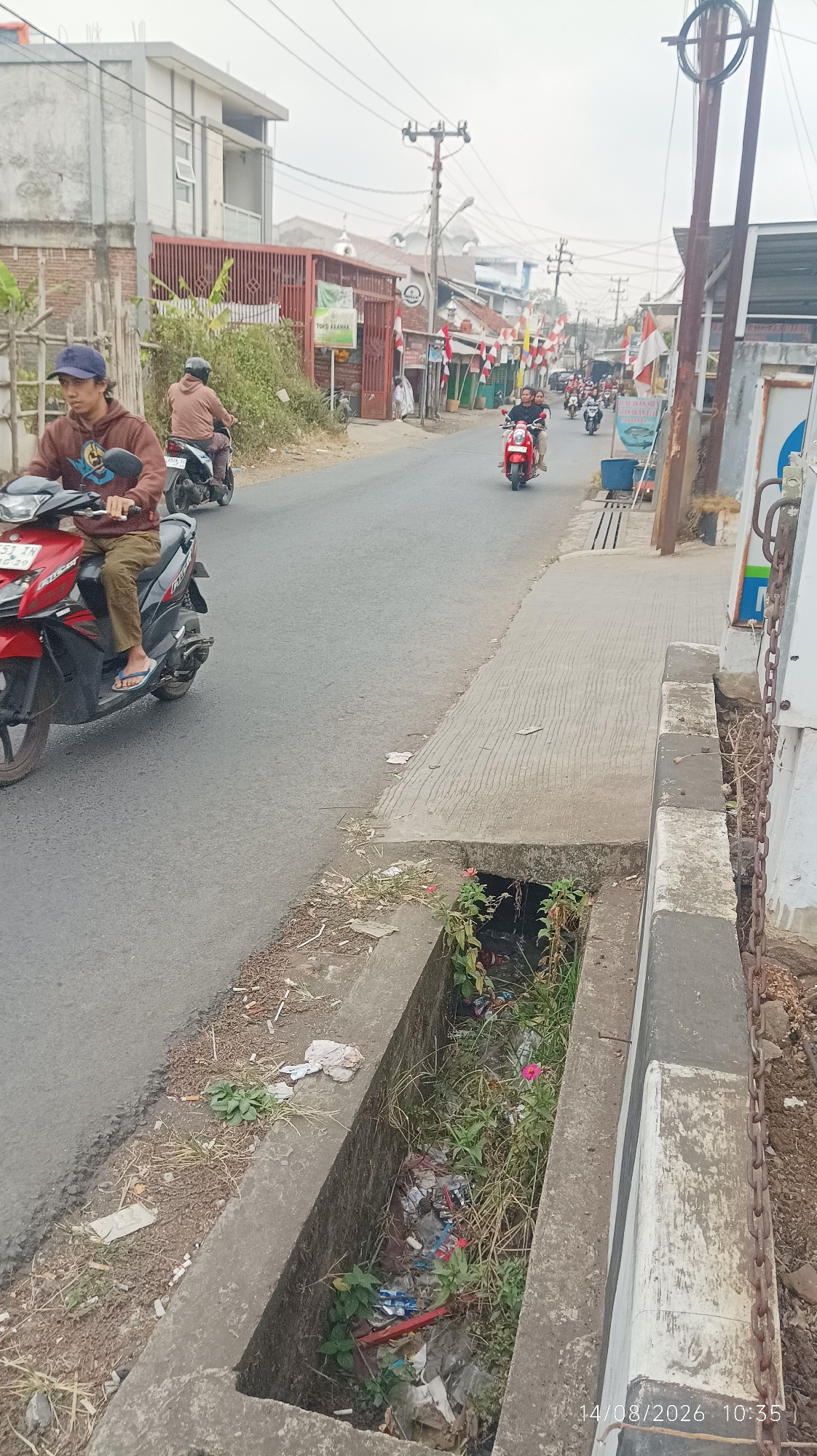 The image depicts a street scene in Tasikmalaya, Indonesia, with a prominent concrete drainage ditch filled with various types of trash, including plastic wrappers, bottles, and other debris. Several people are riding motorcycles on the asphalt road. In the foreground, a young man in a brown hoodie and a blue cap is riding a red and black motorcycle. Behind him, another person on a motorcycle is visible. Further down the road, a red motorcycle with two riders is approaching. The street is lined with buildings, some of which display Indonesian flags, suggesting a festive occasion. Power lines and poles are visible overhead. The environment appears to be a typical urban or semi-urban setting in Indonesia. The date stamp on the image indicates "14/08/2026 10:35".