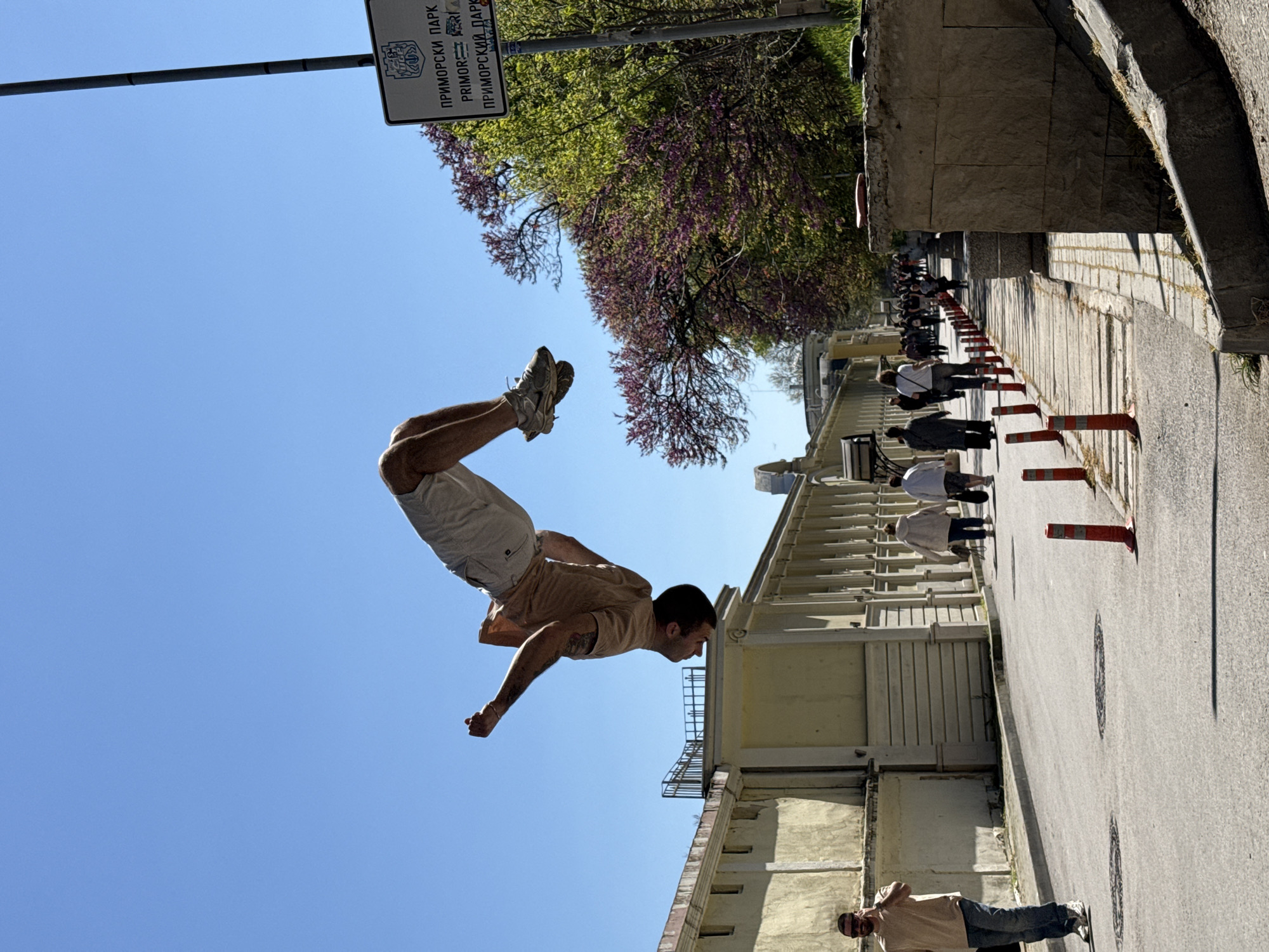 A man is captured mid-air, possibly performing a jump or stunt, in what appears to be a pedestrian area in Varna, Bulgaria. He is wearing a light-colored t-shirt, shorts, and athletic shoes. Below him, another man is standing on the pavement, possibly observing or involved in the stunt. A row of orange traffic cones is lined up along the side of the street, and a group of people can be seen walking in the distance, presumably pedestrians. Buildings line the street, and a sign with Cyrillic text indicates the location is likely near Primorski Park. A tree with purple flowers is also visible. The sky is clear and blue.