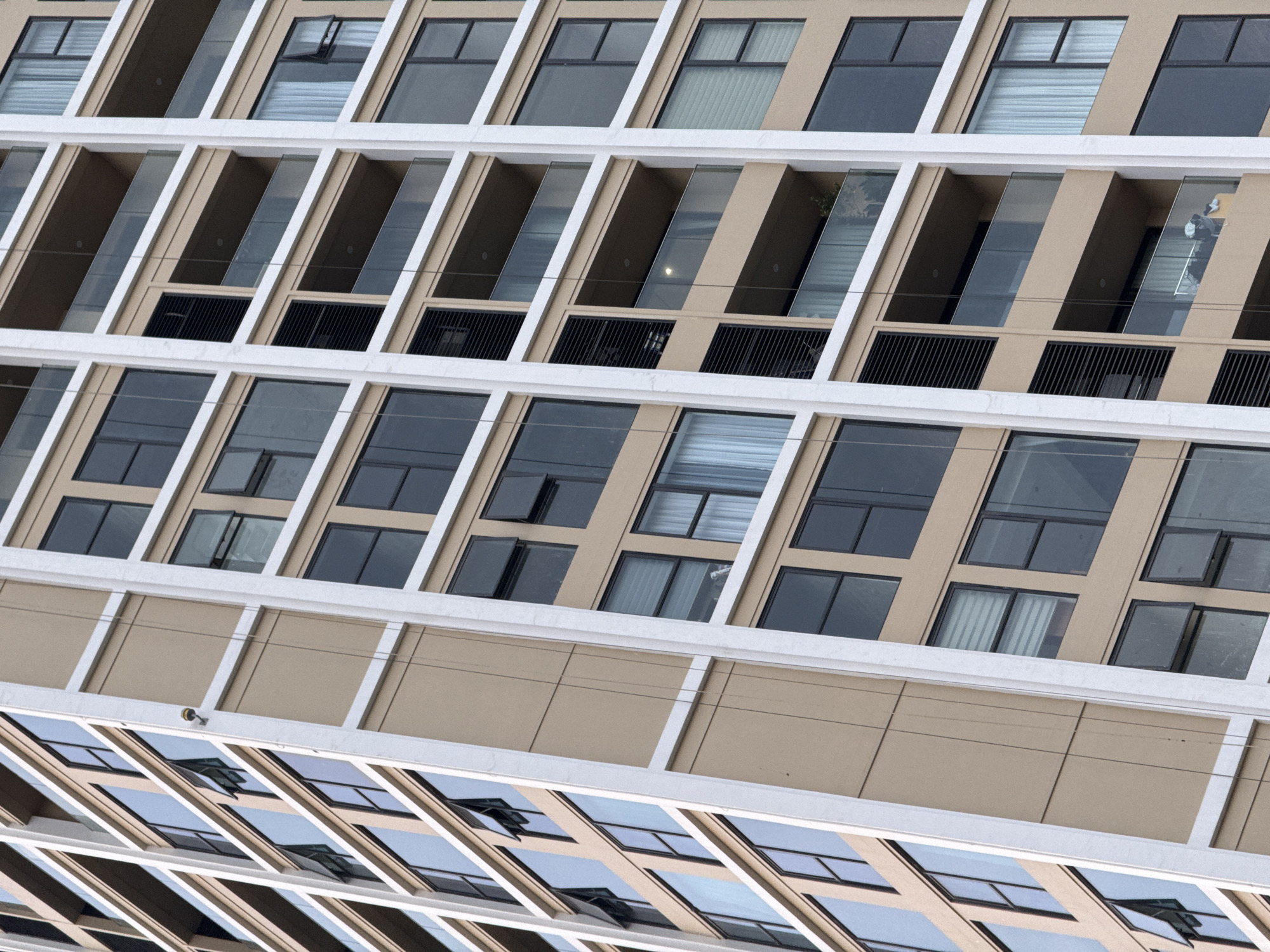 The image shows a close-up, oblique view of the facade of a modern, multi-story building in Da Nang, Vietnam. The building features a grid-like structure of tan-colored panels and white vertical and horizontal framing. Numerous windows, some of which are open, are visible across the various levels. Several balconies with glass railings are also present. A security camera is mounted on the exterior wall of one of the upper floors. The architectural style suggests a residential or commercial building, likely an apartment complex or hotel.