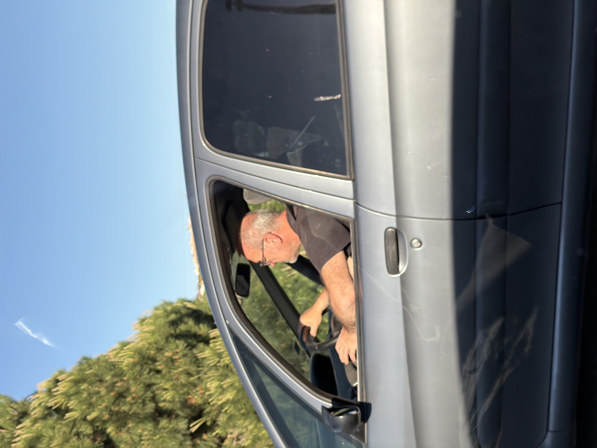 A mature, light-skinned male with short gray hair, glasses, and a dark-colored shirt is shown seated inside the driver's seat of a silver-colored vehicle. The man has his hands on the steering wheel, as if driving the car. The vehicle is parked next to a dense green tree. The location is Mataró, Spain.