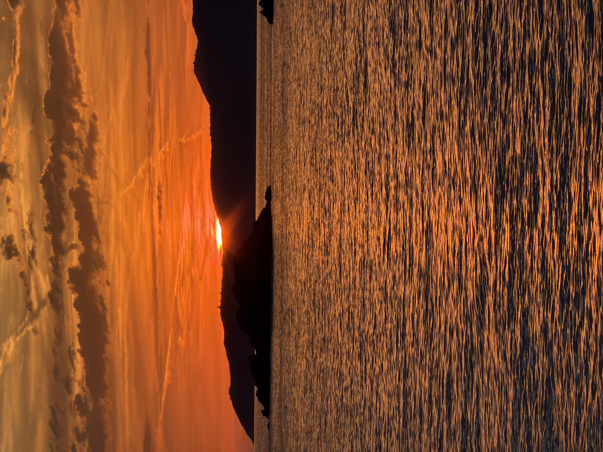 The image captures a sunset scene over the Adriatic Sea near Perazića Do, Montenegro. The sky is dominated by intense hues of orange and red, with scattered dark clouds and faint contrails visible in the upper portion. The sun, partially obscured by a distant, silhouetted mountain range on the horizon, emits distinct rays of light upward and to the right. Several dark, irregular rock formations or small islands are silhouetted in the mid-ground water. The surface of the sea exhibits numerous small, horizontal ripples, reflecting the warm orange light from the sky. On the peak of one of the silhouetted mountains, a slender, dark structure, possibly a communication tower or mast, is discernible. To the far left, a lower, more distant landmass is also silhouetted against the bright sky, showing a fainter structure on its crest. No people or boats are explicitly visible.
