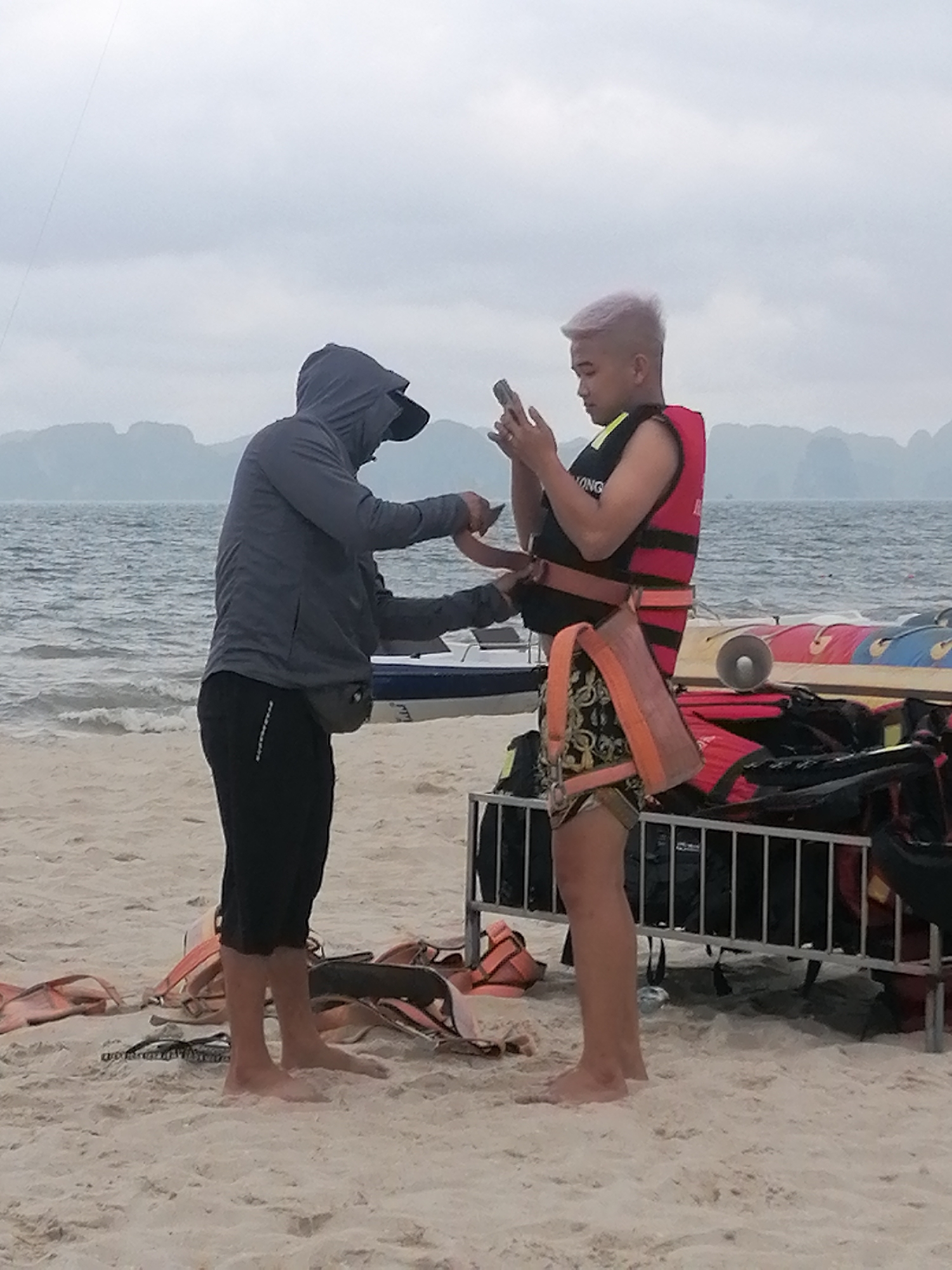 The image captures two men on a sandy beach against a backdrop of a calm, choppy sea and distant karsts under an overcast sky. In the foreground, numerous orange life jackets are scattered on the sand and piled on a metal rack.
On the left, a man wearing a dark grey hooded jacket, black pants, and a fanny pack is standing barefoot. He is leaning towards the other man, seemingly adjusting a strap or harness around his waist. He is also holding a phone in his right hand.
On the right, another man, also barefoot, wears a red life jacket with a partially visible text "LONG" on it, and patterned shorts. He has light-colored, possibly bleached hair. He is holding a smartphone in both hands and looking down at its screen, seemingly engaged with it while being attended to. An orange harness with straps hangs from his waist.
In the mid-ground, a small white boat is partially visible in the water, and behind the men, other water activity equipment, including what appears to be part of an inflatable banana boat or similar, is stacked on or near the metal rack. The overall scene suggests preparation for water-based activities at a coastal location like Bai Chay Ward in Vietnam. The sky is uniformly cloudy, indicating daytime with diffused light.