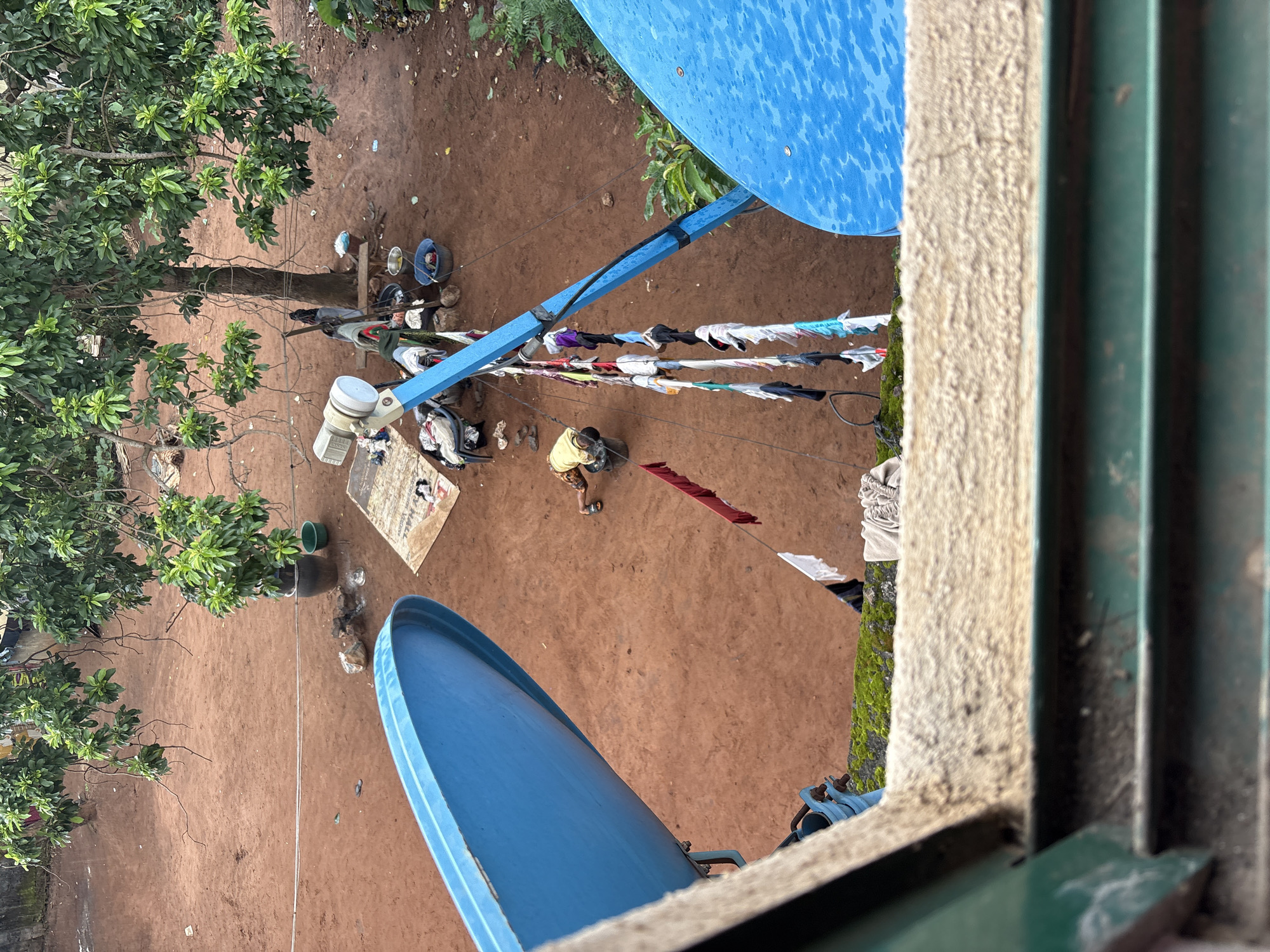 An elevated, slightly angled, full shot depicts a rural or semi-rural scene in Onitsha, Nigeria. A dirt ground fills the middle of the frame, dotted with debris and a few scattered items. In the foreground, a large, bright blue satellite dish occupies the lower left quadrant, partially obscuring the view of the scene beyond. Another blue satellite dish is visible on the right, further into the background.
Dominating the upper portion of the frame is a leafy green tree with a thick trunk, its branches reaching across the space. Beneath the tree, several people are engaged in various activities. A person wearing a yellow shirt and dark shorts is bent over, appearing to be working with a dark bucket. Nearby, a collection of clothes hangs on a clothesline, some colorful and others white, suggesting laundry is being air-dried. A blue satellite dish mount or pole extends vertically through the scene, supporting the clothesline.
Other objects in the scene include buckets, what appear to be chairs, and scattered papers or fabric. The ground is a reddish-brown earth, with some areas appearing dry and dusty, while others might be slightly damp. The overall impression is one of daily life unfolding in an outdoor setting.