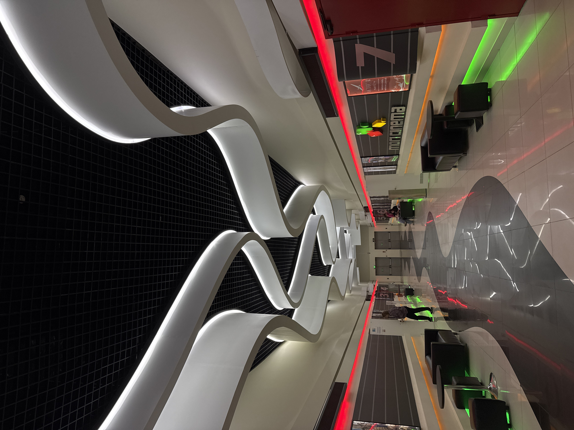 The image shows the interior of a modern cinema, possibly in a shopping mall, located in the city of Unknown, Unknown. The camera is positioned low to the ground and at an angle, emphasizing the perspective.
The architectural design is characterized by long, curved white structures that appear to be suspended from the ceiling. These structures are lined with lights, creating a bright, modern aesthetic. A black grid pattern is visible behind the white structures.
Along the right side of the image are signs indicating the cinema halls, marked with numbers and lit with red and green neon. A row of black lounge chairs with small tables are positioned next to the hall signs, suggesting a waiting area. People are visible in the background, walking towards or waiting near the cinema halls.
The floor is polished and reflects the lights and colors of the environment, creating a dynamic visual effect. The overall impression is a sleek, contemporary, and well-lit public space.