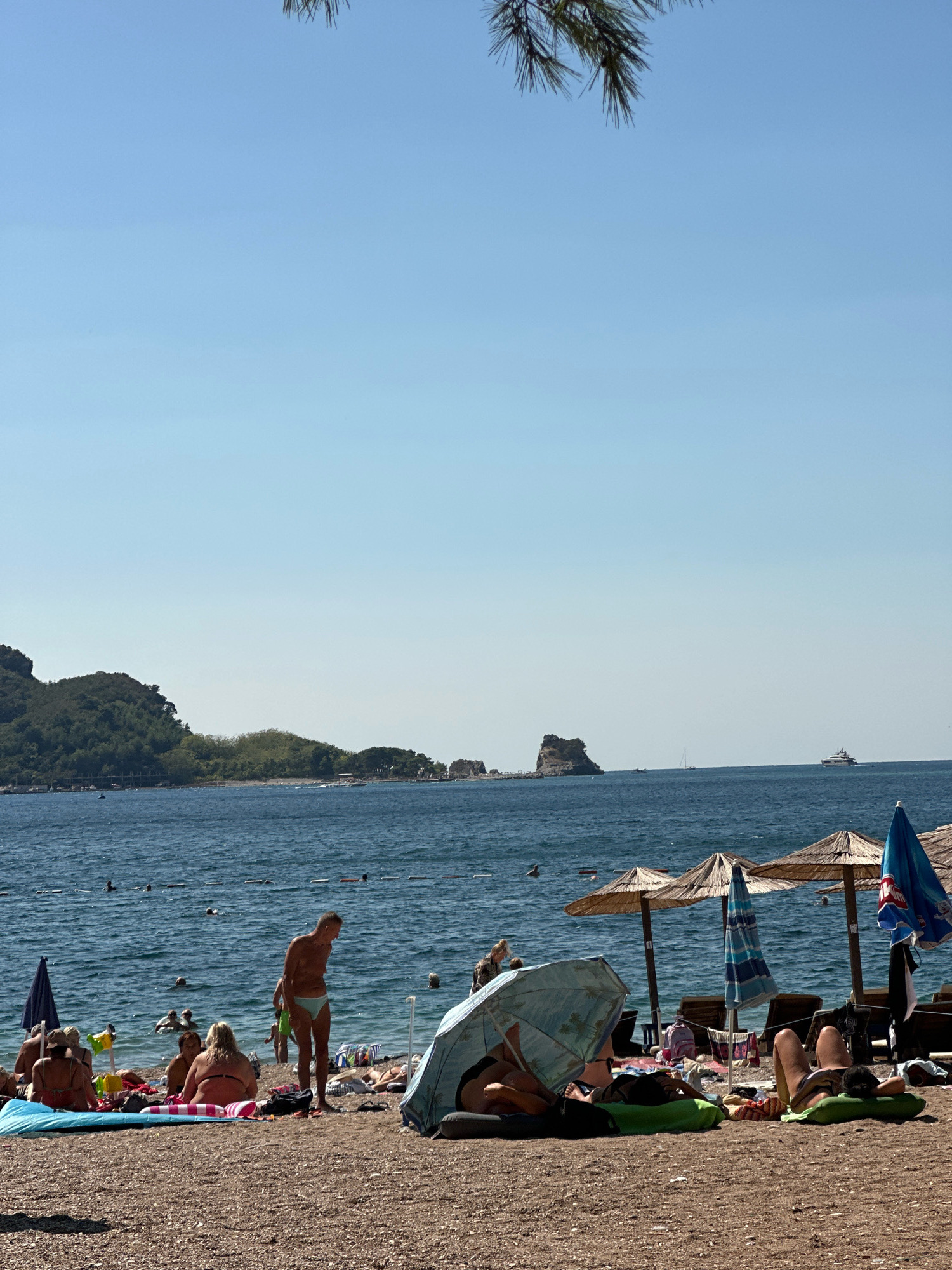 The image depicts a sun-drenched beach scene in Budva, Montenegro, with a clear blue sky overhead and a gentle sea breeze. The beach is covered in small pebbles. In the foreground, several people are sunbathing on towels and loungers. A man in light blue swim trunks stands, looking down, while a woman in a bikini sits beside him. To the right, two individuals recline under a patterned umbrella. Further back, more beachgoers are visible, some in the water.
The sea is a deep blue, with a few distant boats on the horizon. In the middle ground, a rocky island with some vegetation rises from the water, and further to the right, a large yacht is visible. The coastline in the background is lined with green hills and trees. Beach umbrellas and loungers are scattered across the sand, some occupied by people relaxing. The overall atmosphere is one of leisure and summer vacation.