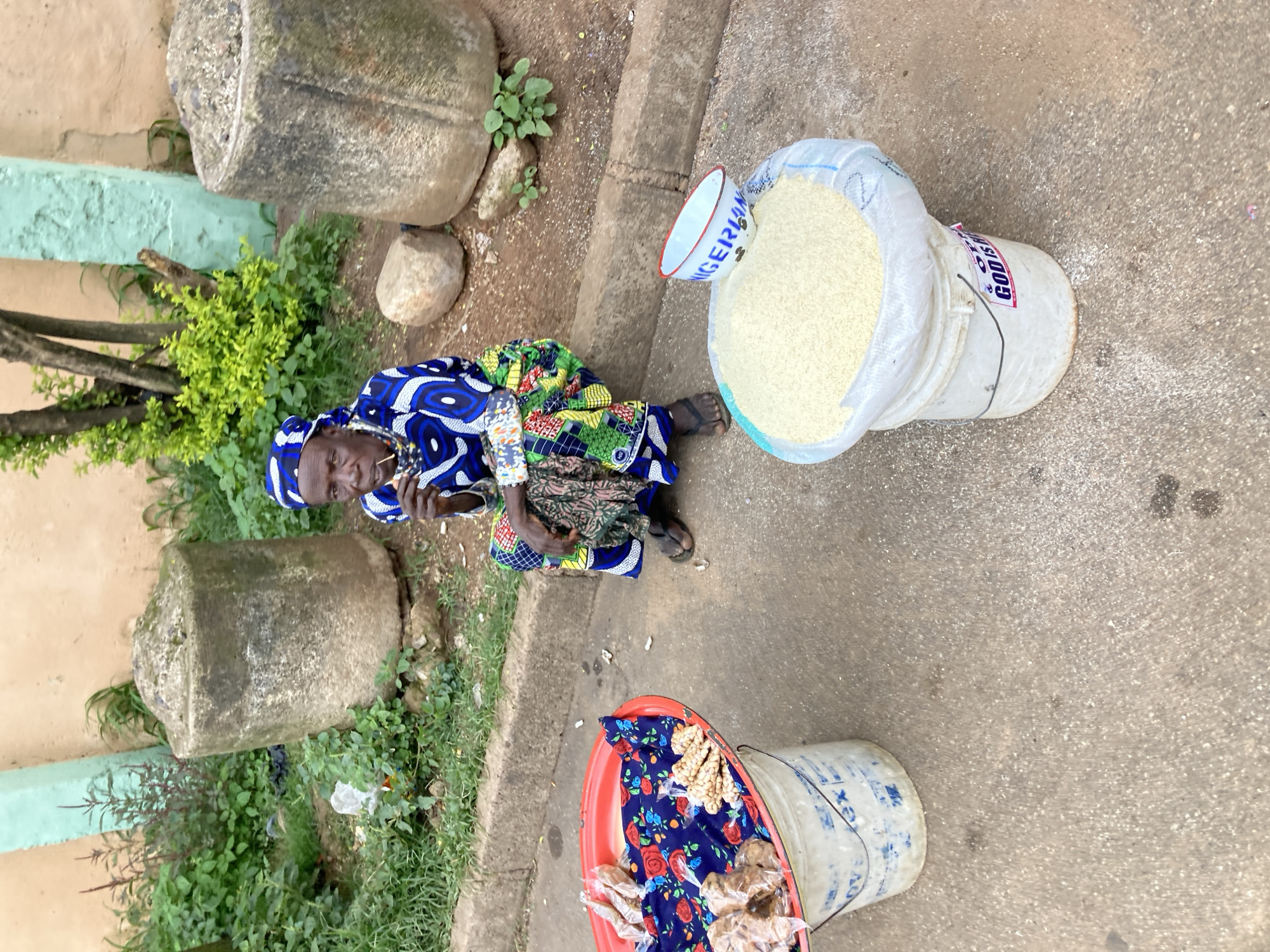 The image shows a woman, likely a vendor, sitting on the ground in an outdoor setting. She is wearing a vibrant blue, white, and green patterned fabric wrap and headscarf. In front of her, a large white sack is filled with what appears to be rice. A white bowl with "NIGERIAN" written in blue lettering is placed on top of the sack. To her right, a red plastic basin is filled with packaged snacks, possibly peanuts. The ground is made of concrete or a similar paved surface, with some patches of dirt and grass. In the background, there are large, weathered stone objects and some greenery. The scene suggests a marketplace or a roadside vending area. The lighting appears to be natural daylight, with no strong shadows, indicating it could be daytime, possibly morning or afternoon. The overall atmosphere seems to be that of daily commerce in a Nigerian locale. There is also some visible text on the sack and the bowl. The text on the sack is partially obscured but includes what looks like "GOD IS."