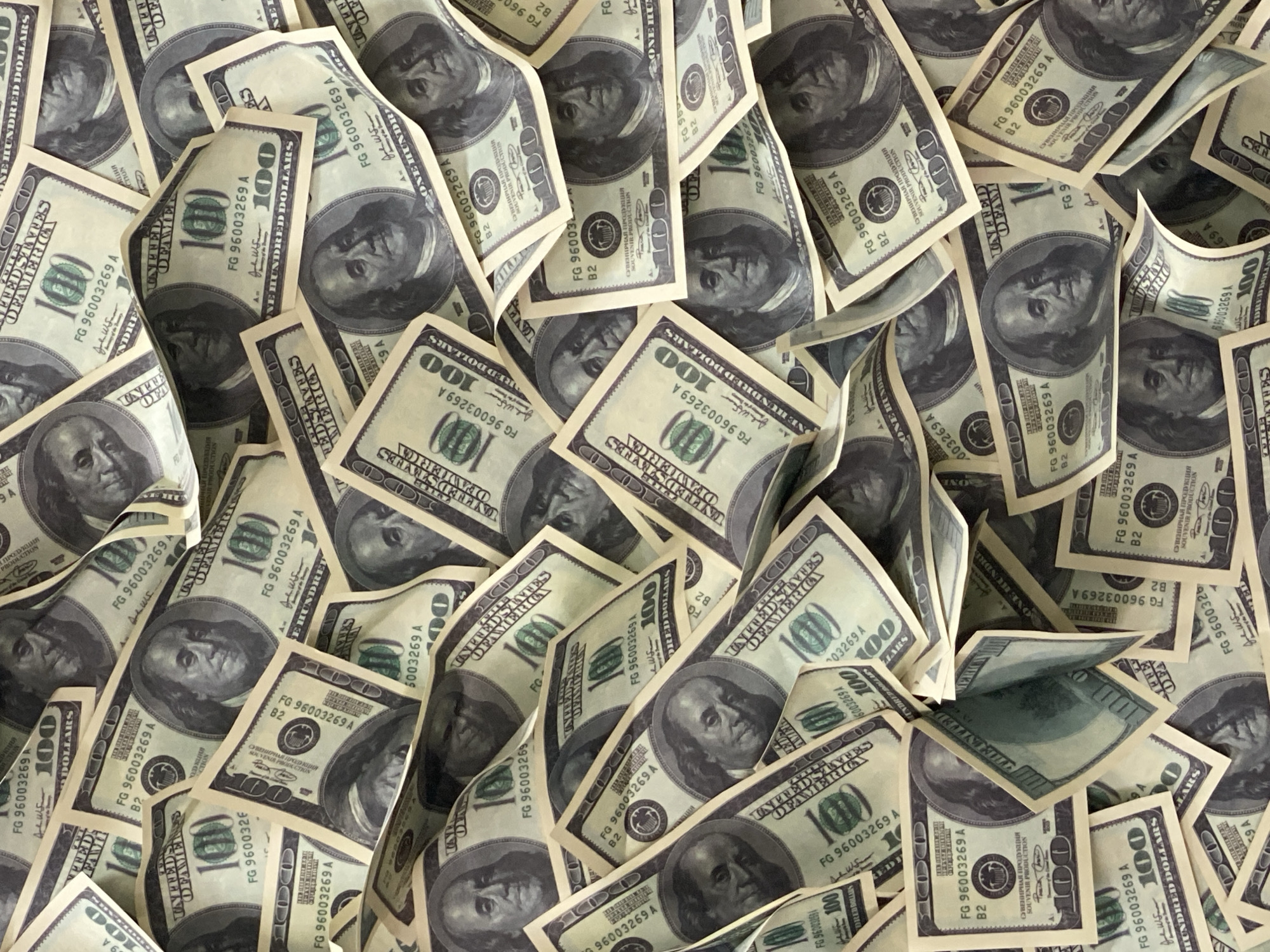 The image shows a large pile of what appear to be stylized, or souvenir, 100 dollar bills. The bills are not real currency. They are scattered and overlapping, creating a dense, textured pattern. The bills feature a portrait, presumably of Benjamin Franklin. The text on the bills includes the serial number "FG 96003269 A" and phrases in Russian, such as "СУВЕНИРНАЯ ПРОДУКЦИЯ", which translates to "Souvenir Production". There is no indication this image was captured in the city of Sochi, Russia.