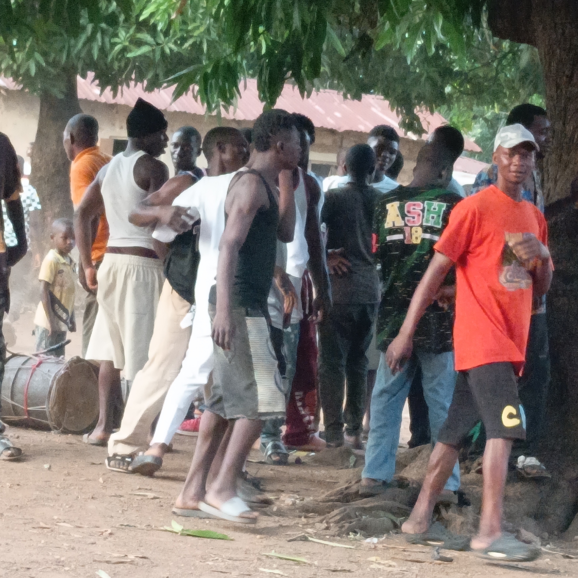 In Akwanga, Nigeria, a group of people congregates outdoors near a tree. A fair-skinned drum lies on the ground next to a child. Men wear a mix of tank tops, t-shirts, shorts, trousers, and sandals. One man stands out, wearing an orange t-shirt and a white baseball cap. The surroundings suggest a casual, informal setting.