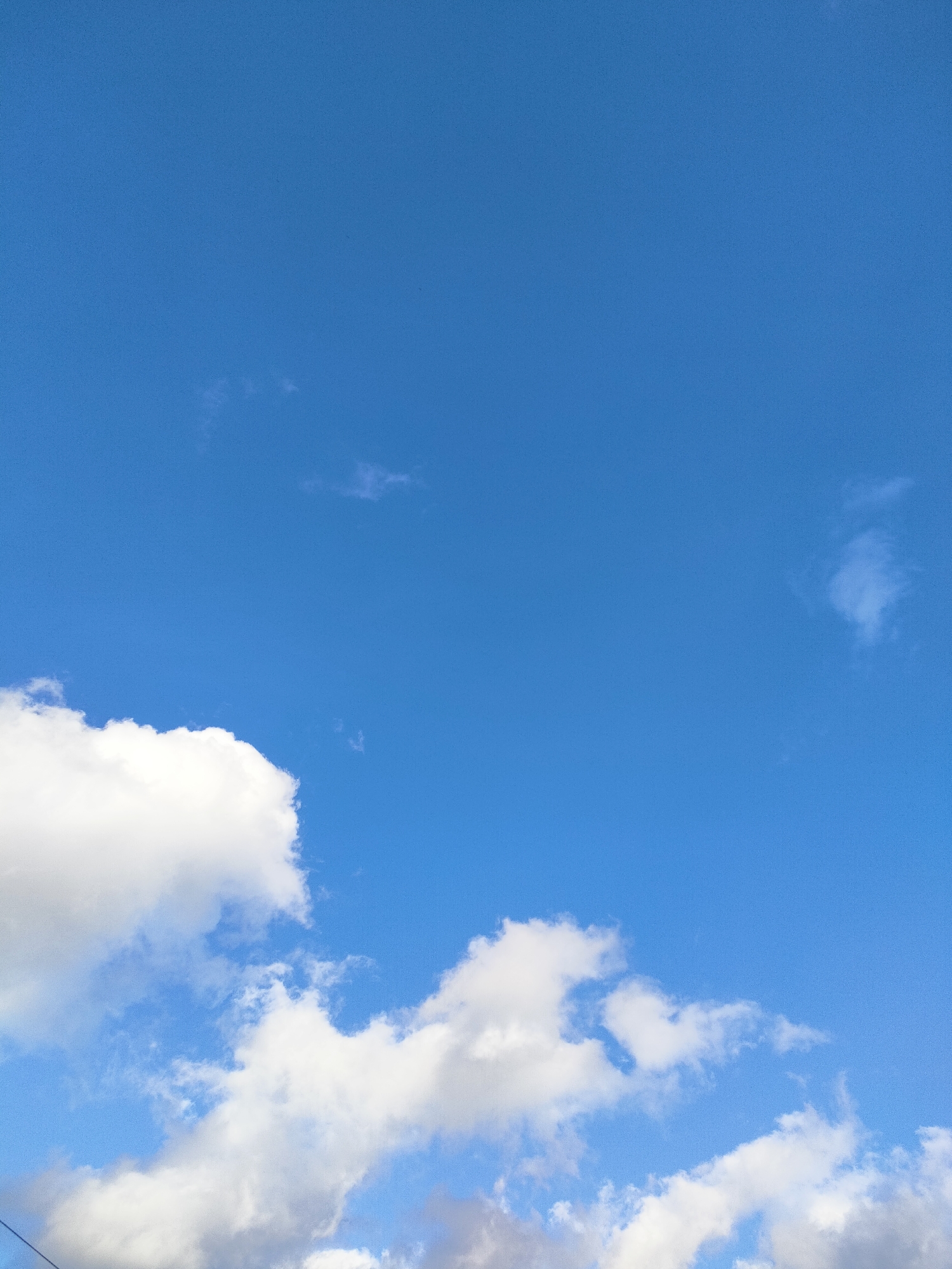 The image is a vertical, medium-shot photograph of a bright blue sky with scattered white clouds. The lower portion of the frame is dominated by a large, fluffy cumulus cloud on the left, and a cluster of smaller clouds to its right. In the upper portion of the sky, there are a few wispy, thin clouds. The lighting suggests it is daytime, with the sun likely high in the sky, illuminating the clouds and giving the sky its vibrant blue hue. There are no people or objects visible in the frame, and no text is present. The scene evokes a sense of openness and a pleasant, clear weather day.
