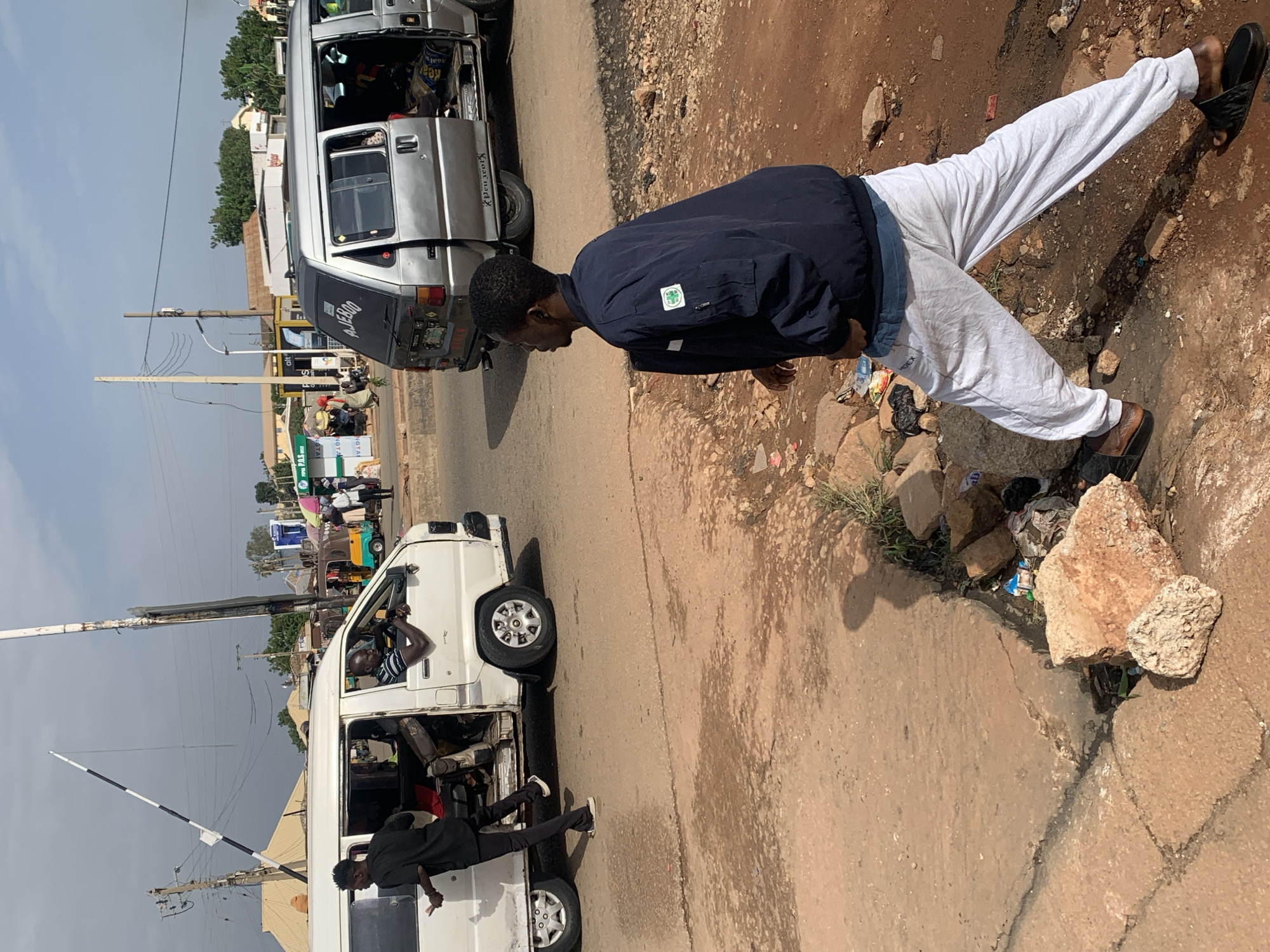 In Gura Top, Nigeria, a bustling street scene unfolds. A white minibus, its passenger door open, is parked on the right side of the road. A man in a black shirt and pants stands outside the minibus, facing forward. Inside, a man in a striped shirt is visible behind the steering wheel. To the right, a silver minibus with its rear door open is also parked. The word "AJERO" is visible on its back.
A young man in a navy jacket and light grey sweatpants walks away from the camera, his back to the viewer. He wears dark sandals on his feet. The road surface is cracked and dusty, with debris and rocks scattered along the side. In the background, buildings and various forms of transportation, including what appears to be a train crossing barrier, suggest an active urban environment. The sky is overcast with some visible power lines.