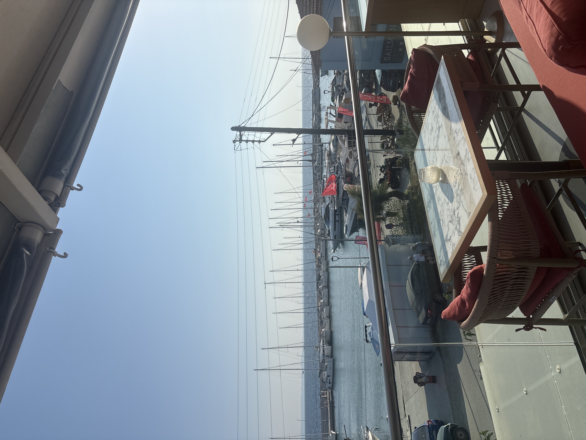 The image captures a sunlit view from a balcony overlooking a marina in Sithonia Municipal Unit, Greece. In the foreground, a marble-topped table with a glass candle holder is set with two chairs, each featuring red cushions. Beyond the balcony railing, a row of sailboats and motorboats are moored in the clear blue water. The marina is lined with docks, and a paved walkway runs alongside it. On the walkway, a few cars are parked, and a red ATV is visible. In the background, power lines stretch across the sky, and a distant landmass can be seen across the sea. The overall scene conveys a sense of leisure and a Mediterranean coastal atmosphere.