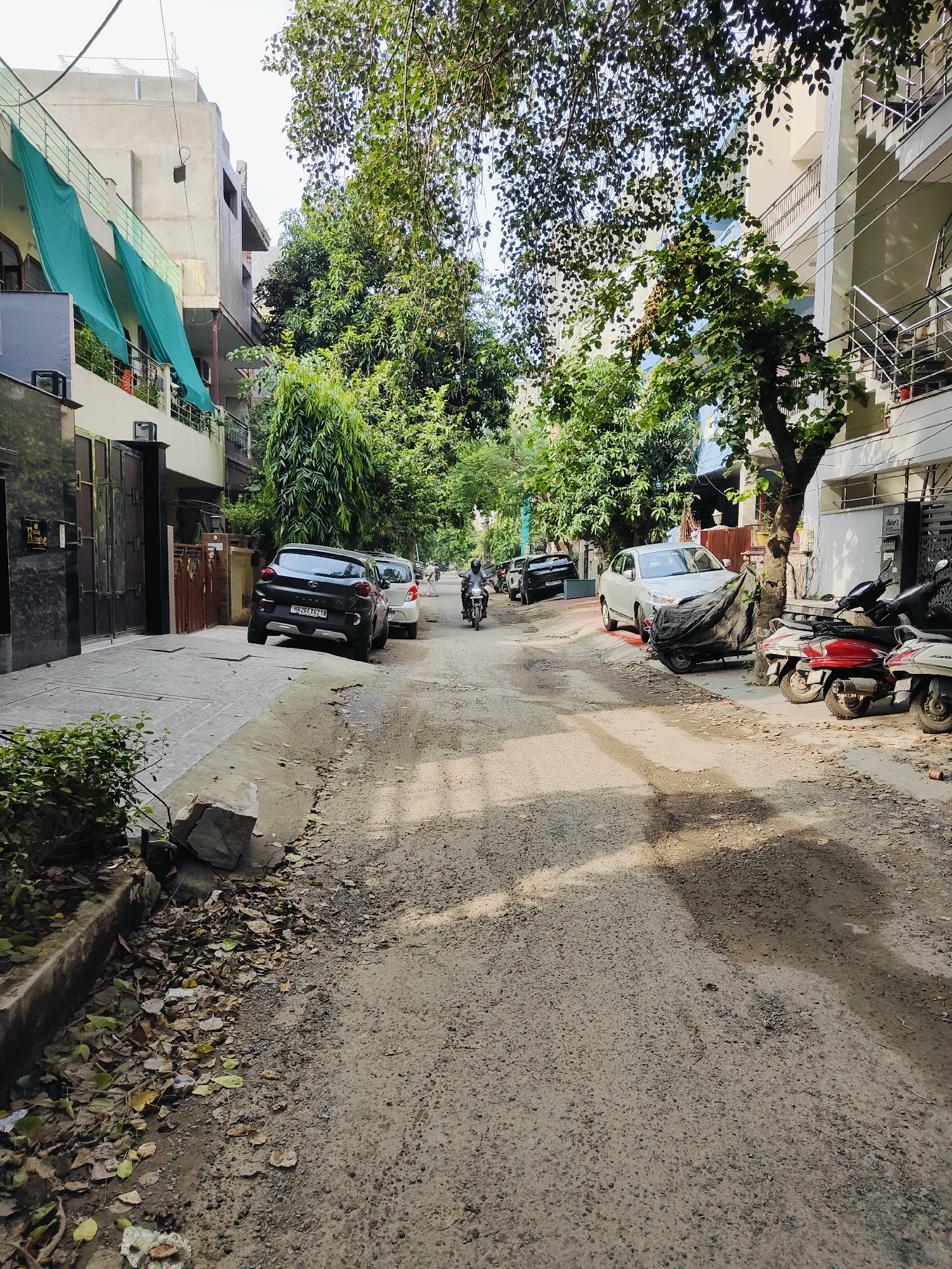 The image depicts a street scene in Gurgaon, India, characterized by residential buildings and a moderately paved road. On the left side of the street, a multi-story building with balconies covered by green awnings is visible. Adjacent to it is a dark-colored gate. The road itself appears somewhat uneven, with patches of loose gravel and dirt.
Several vehicles are parked and in motion. A black SUV with license plate HR26EX6210 is parked on the left side of the road, facing away from the camera. A white car is parked behind it, with a black tarp covering a portion of its front. Further down the street, several scooters and motorcycles are parked on the right side. A person on a motorcycle is traveling away from the camera in the middle of the road.
The environment is characterized by the presence of numerous trees lining the street, casting shadows on the road. Fallen leaves are scattered on the side of the road and sidewalk area. The sky is visible at the top of the image, appearing partly cloudy. The overall impression is a typical urban street in a residential area of Gurgaon, India, with elements of everyday life captured in the scene.