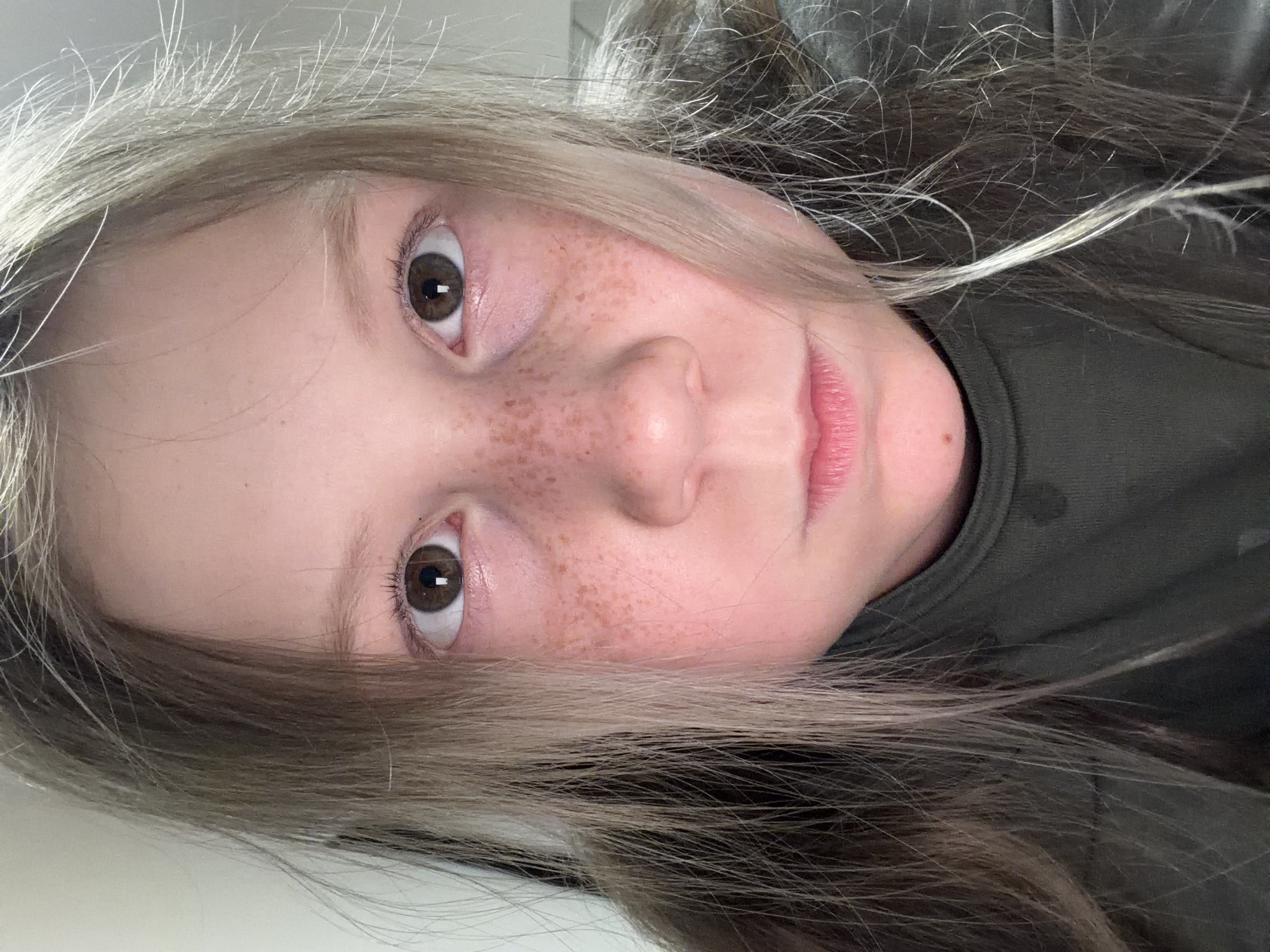 A close-up portrait depicts a young girl with fair skin, brown hair interspersed with lighter blonde strands, and brown eyes. She has freckles across her nose and cheeks. The background consists of a solid, light-colored surface to the left and a dark green or grey fabric on the right side. The image appears to have been taken indoors. There's no indication in the image itself that would reveal a specific location in Markovo, Russia.