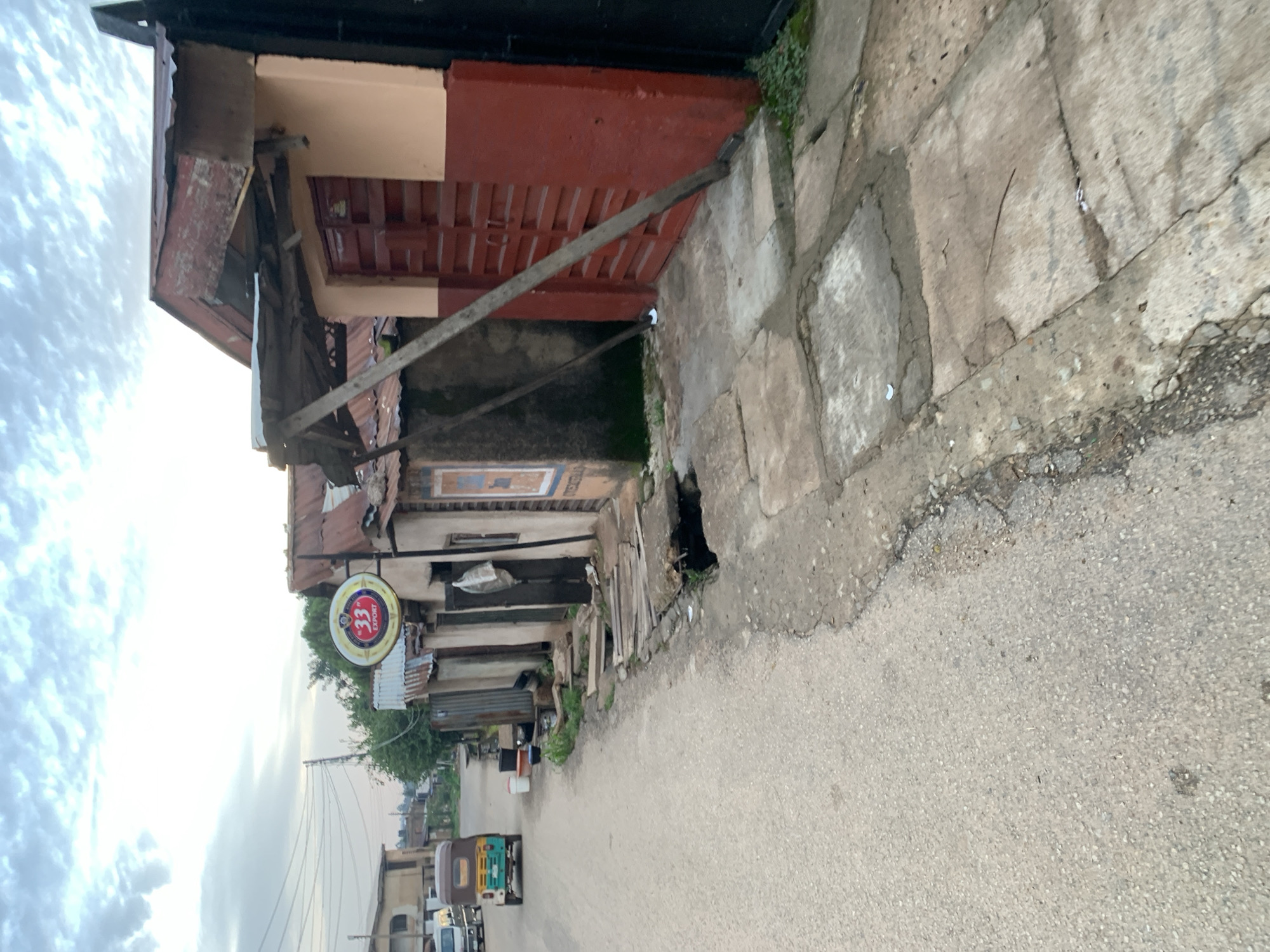 An eye-level, medium shot captures a street scene in Jos, Nigeria, under a cloudy sky. The perspective looks down a paved road on the left, leading toward a small, weathered building on the right. A yellow and green motorized tricycle, a common mode of transport in Nigeria, is parked on the left side of the road. The building on the right features a red-paneled door and a peach-colored upper wall, with a rusted corrugated metal roof. A prominent circular sign for "33 EXPORT" beer hangs above the entrance. The foreground shows a deteriorating concrete sidewalk with visible cracks and a partially open drainage ditch. Evidence of wear and tear is apparent throughout the scene, with corrugated metal and wooden structures forming part of the building's facade and roof. The overall impression is one of an urban environment in a state of disrepair.