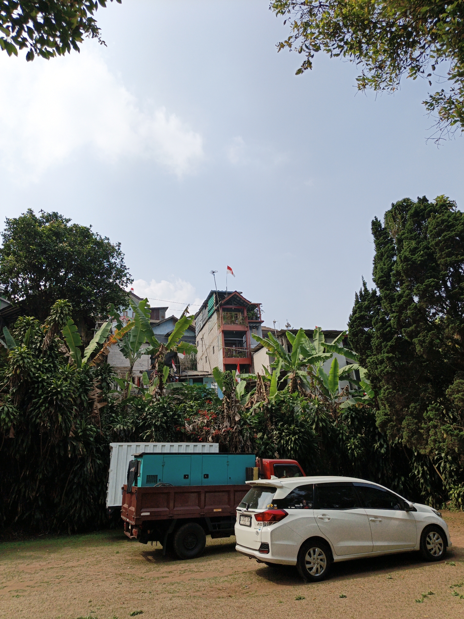 In Cibogo, Indonesia, a white multi-purpose vehicle is parked on a dirt lot, facing right. Behind it, a flatbed truck with a turquoise generator and a white cargo container is parked. Dense green foliage, including banana trees, and trees are abundant in the background, with multi-story buildings visible between the trees, one of which has a red and white flag on its rooftop. The sky is clear with scattered clouds.