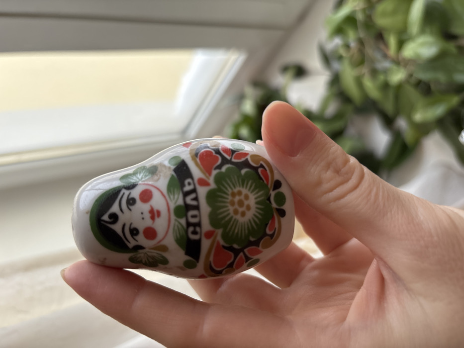A light-skinned human hand holds a white ceramic salt shaker shaped like a traditional Russian matryoshka doll. The doll is adorned with a folk art design featuring a woman's face with black hair, green headscarf accents, red cheeks, and red lips. Below the face, a black band displays the Cyrillic word "СОЛЬ" (SOL), meaning "SALT," in white letters. The lower body of the shaker exhibits a large green floral pattern with red and green embellishments. The scene is illuminated by natural light entering through a window with a white frame in the background. Beyond the window, a light-yellow exterior is out of focus. Blurry green foliage is visible to the lower right. The specific location of this scene is in the city of Velikiye Luki, Russia.